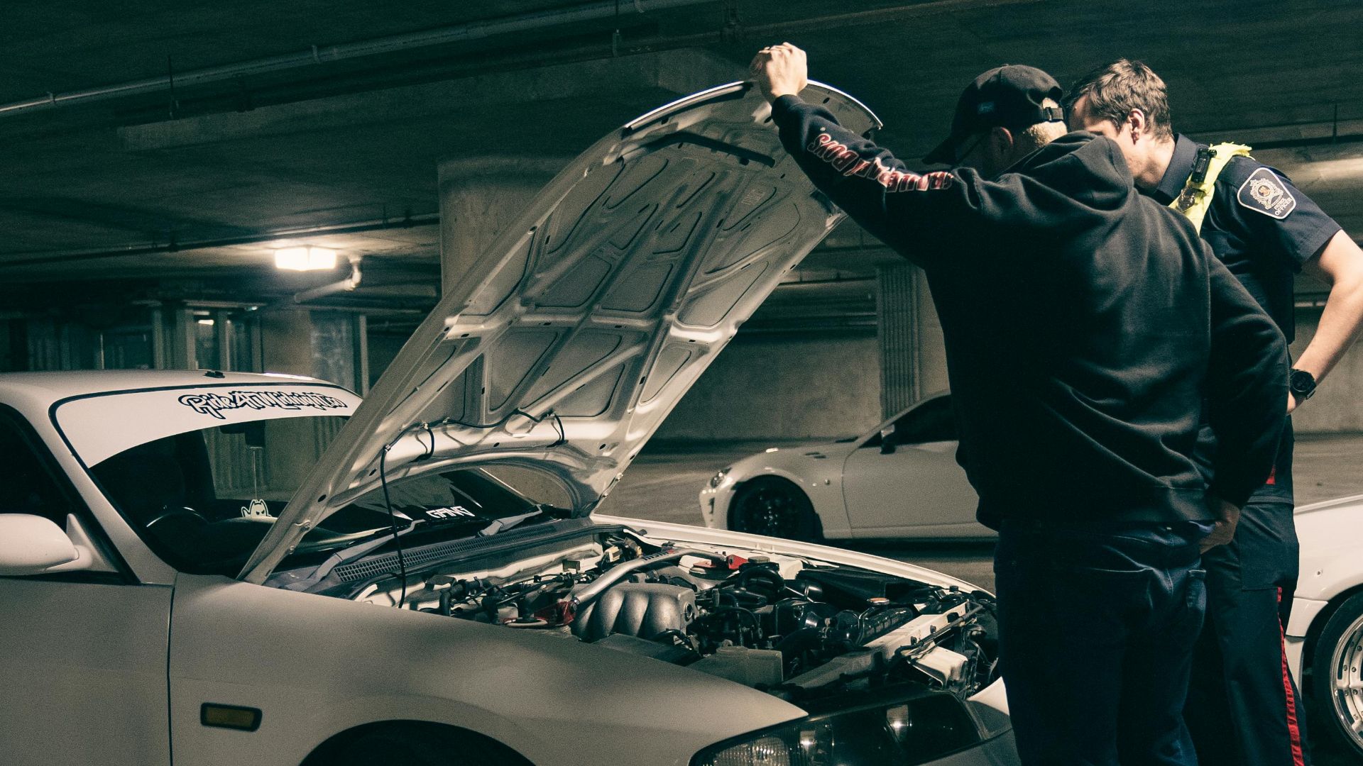 Two men examining a car engine in a dimly lit underground parking area.