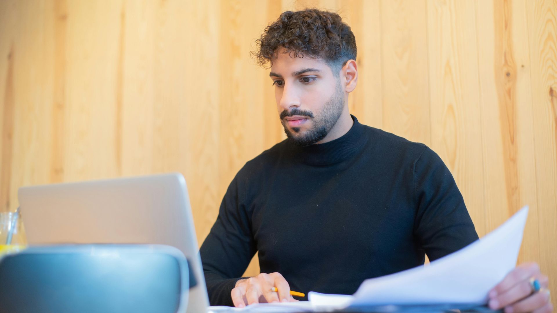 Focused businessman with beard working on a laptop, reviewing documents in a modern workspace.