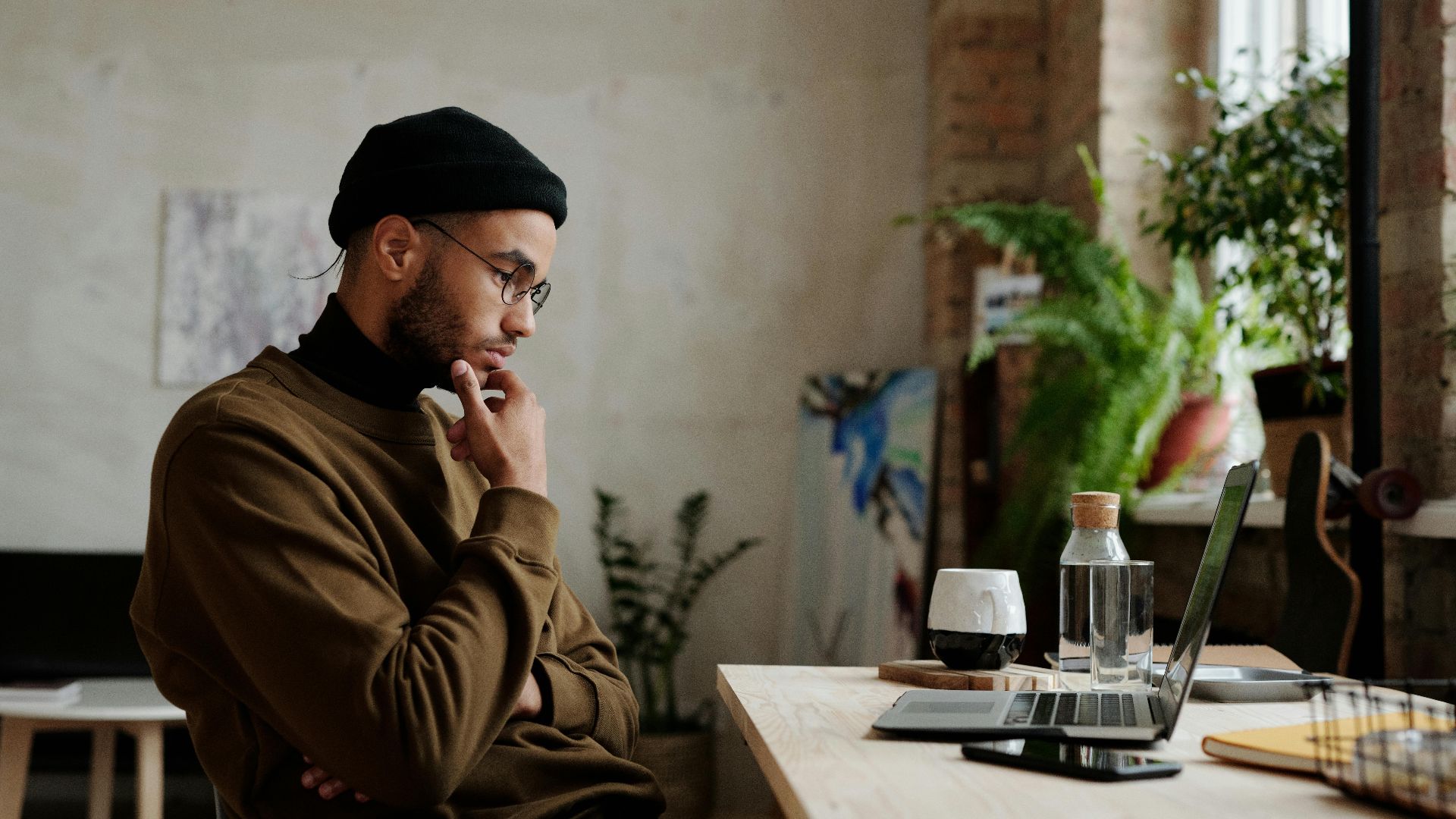 A man in casual attire working thoughtfully on a laptop in a cozy home setting with plants.