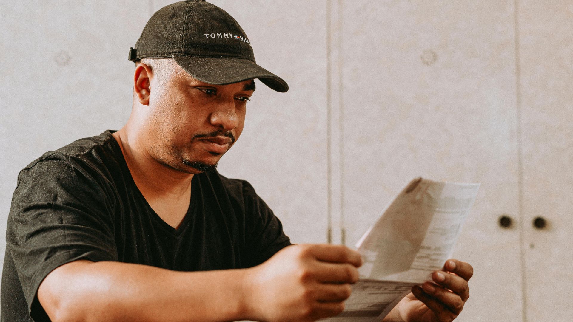 A man in a black cap reading papers at a table indoors.