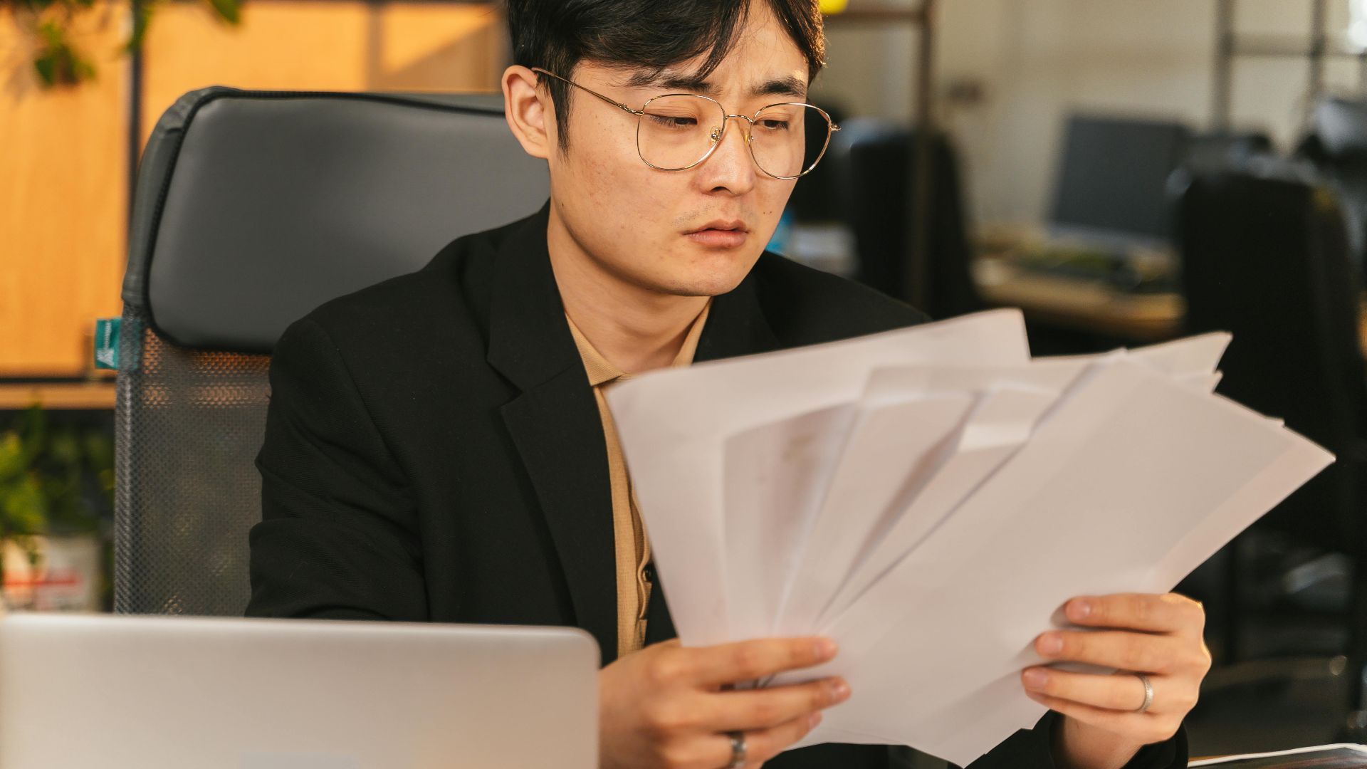 Asian man in glasses and suit analyzing papers at his desk in a modern office.