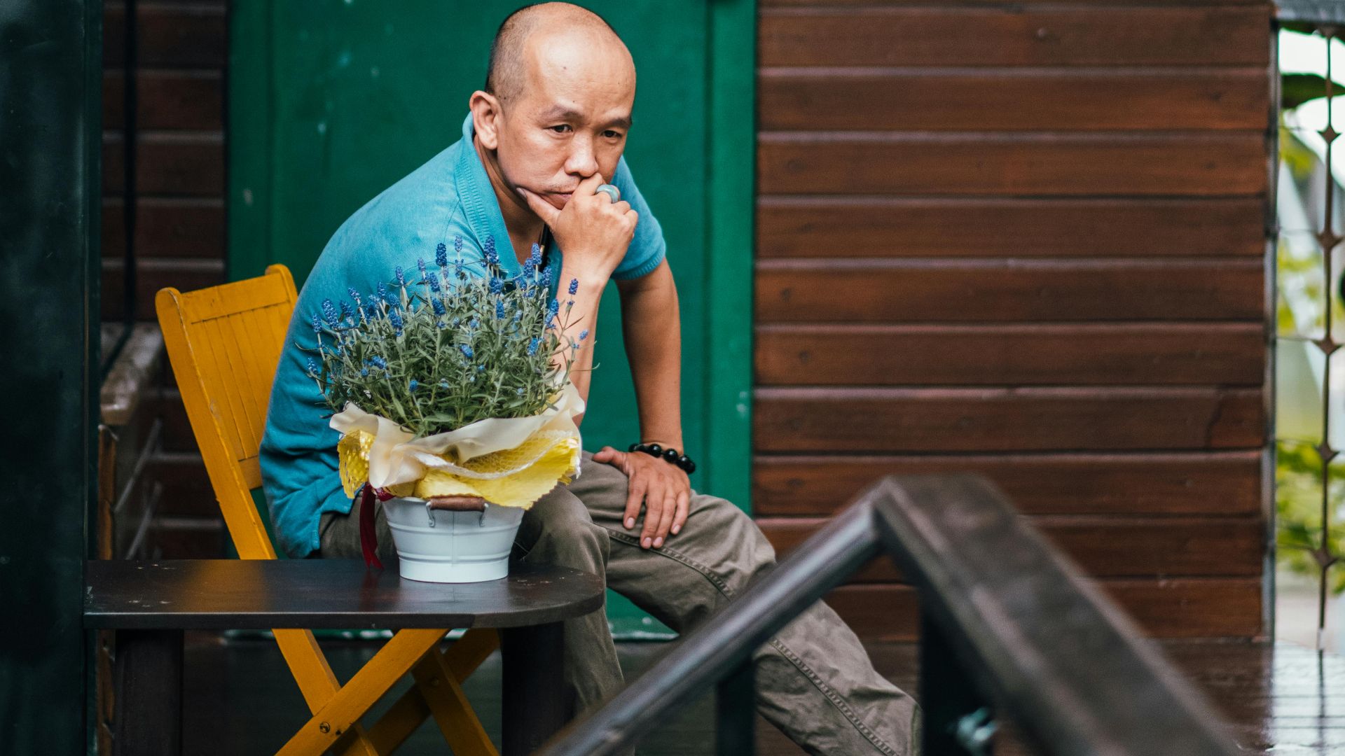 Bald man in casual shirt seated outdoors by a wooden cabin, contemplating.