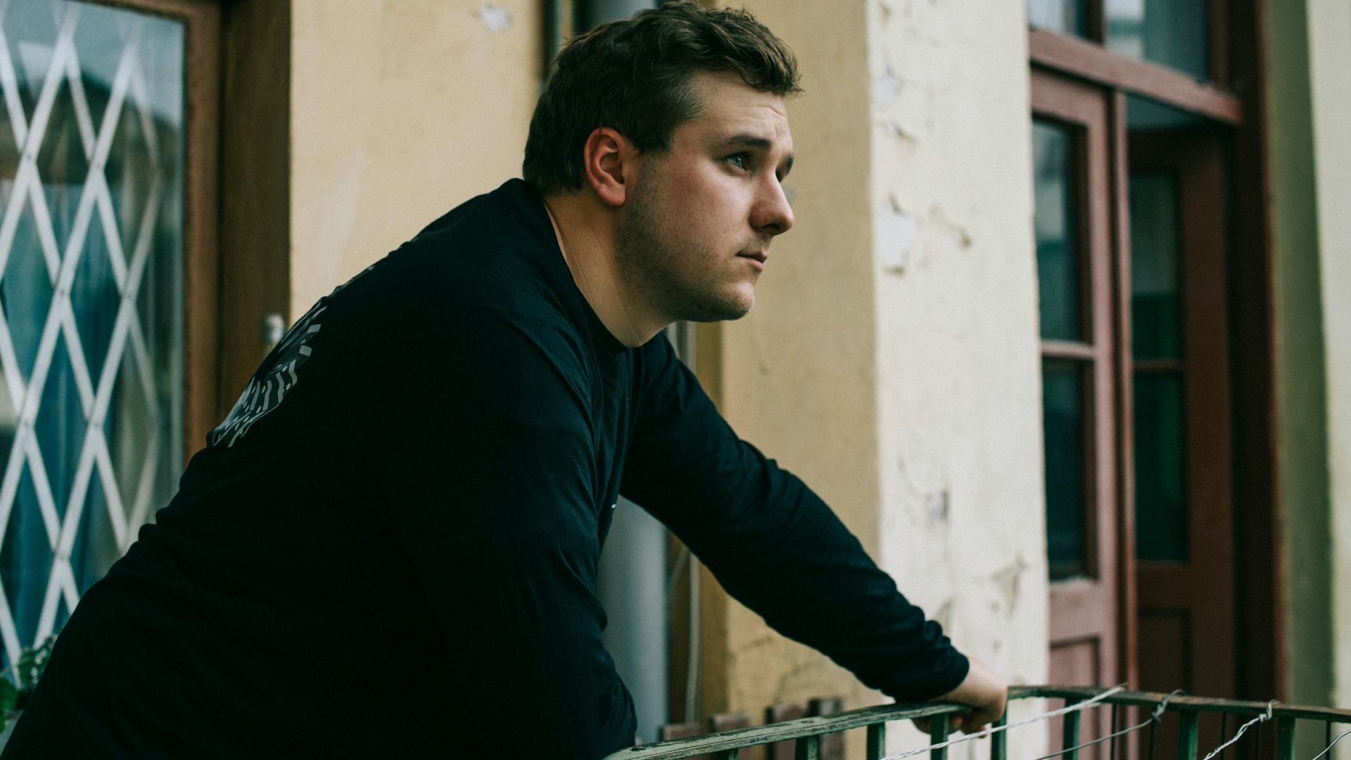 A man in casual attire leans on a balcony, gazing thoughtfully. Urban setting, architectural details.