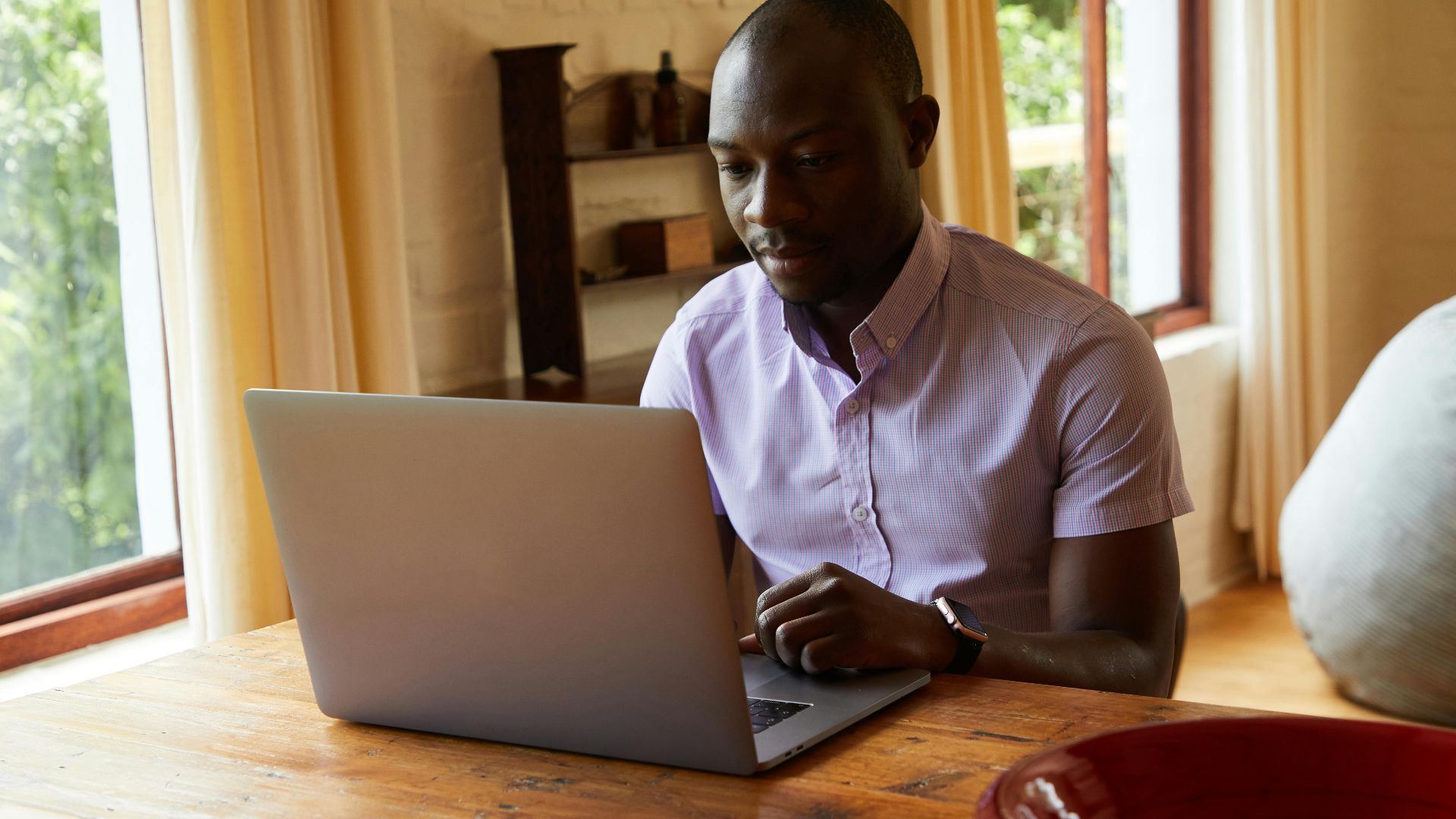 Serious African American male in shirt surfing internet on netbook placed on timber table in comfortable room
