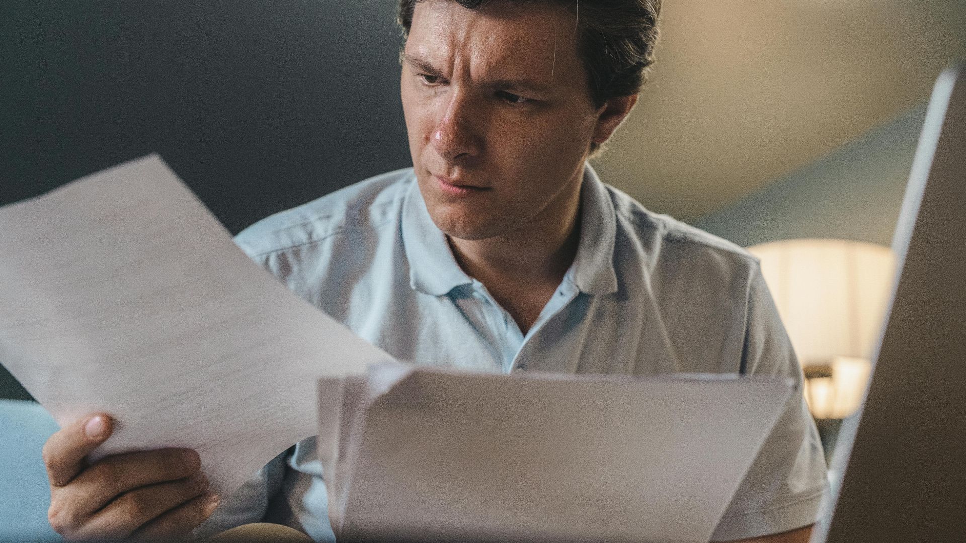 A man intently examines papers, seated indoors under warm lighting, focusing on his work.