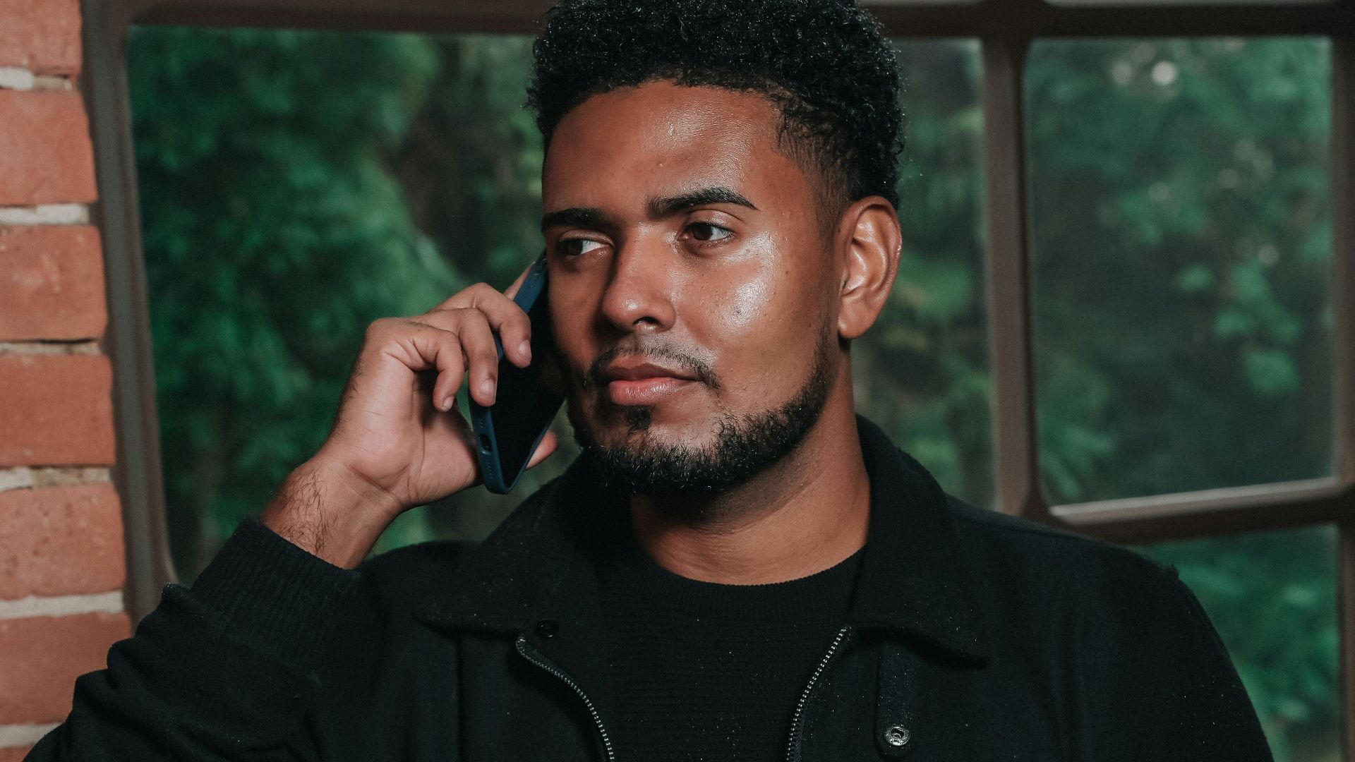 Young man talking on phone by a large window with brick walls.