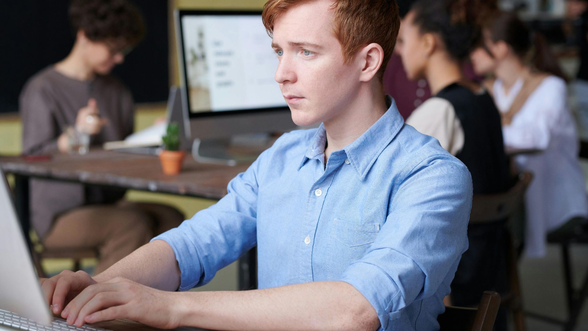 Young professional working intently on a computer in a busy modern office setting, surrounded by colleagues.