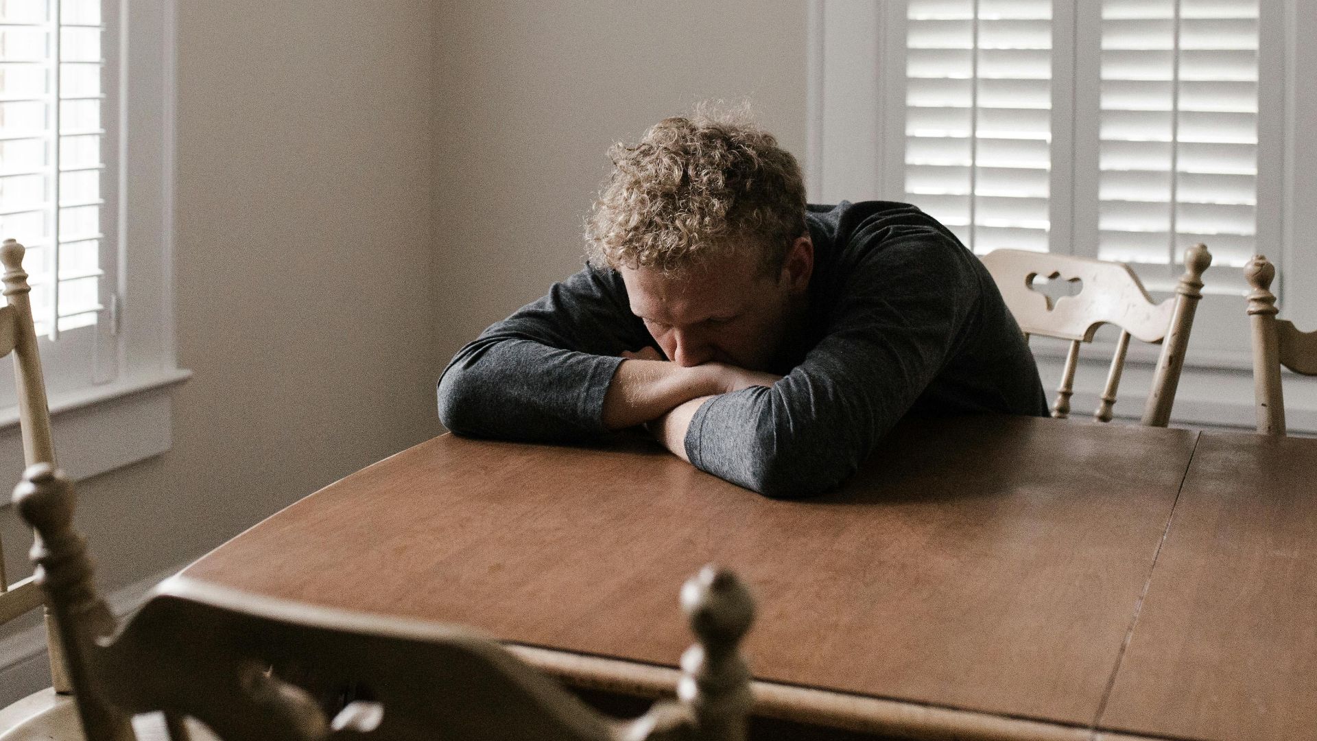 An adult man with emotions of sadness and anxiety sitting alone at a wooden table near windows.