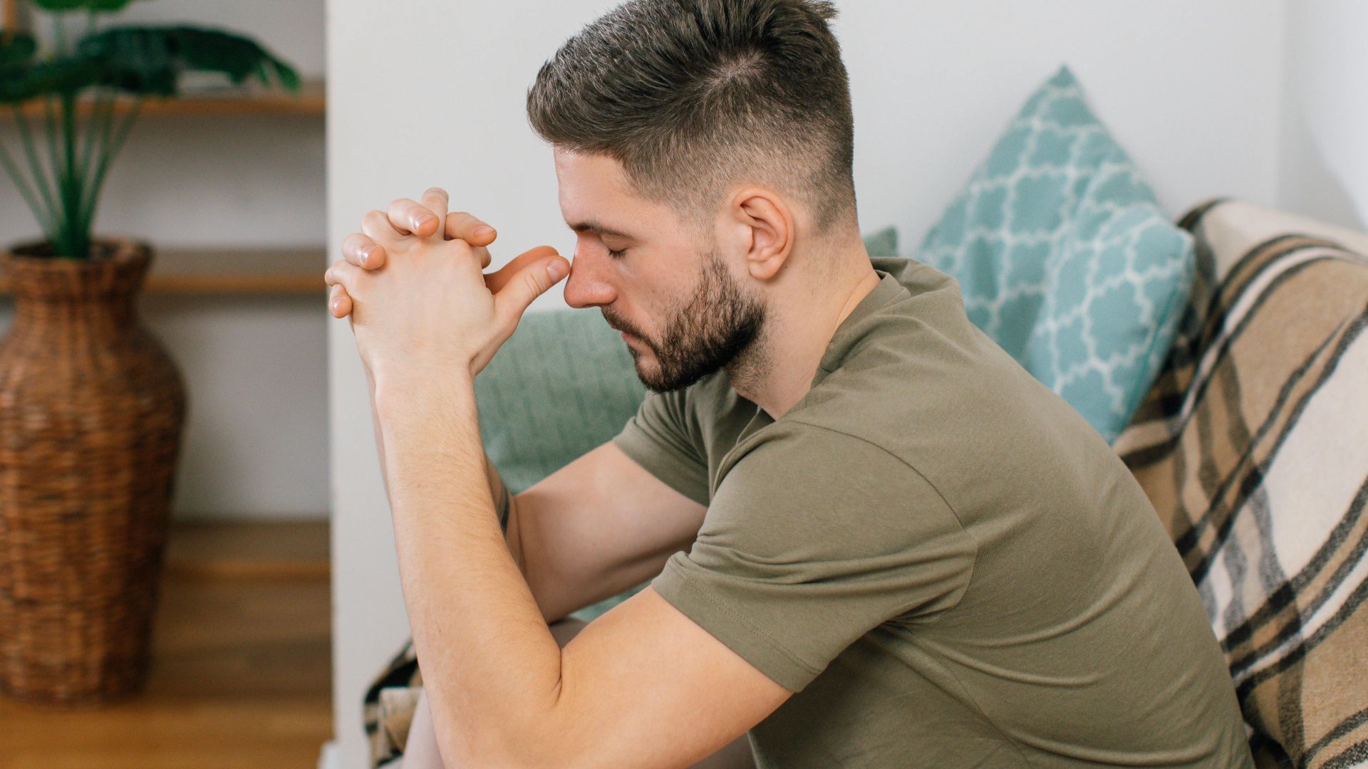 A young man with clasped hands, sitting thoughtfully on a sofa in a cozy living room.