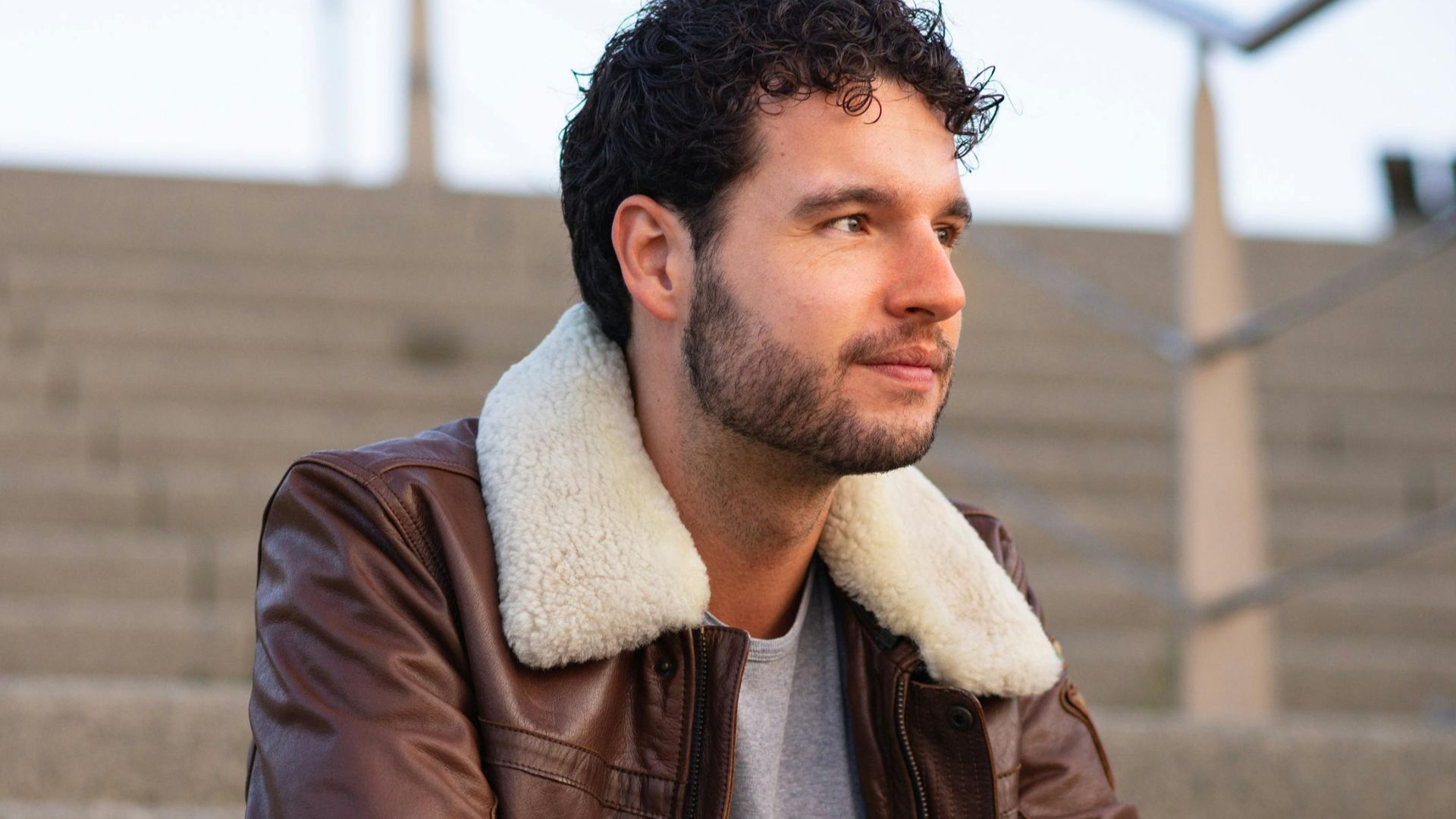 A young man gazes thoughtfully while seated on urban steps, wearing a stylish leather jacket.