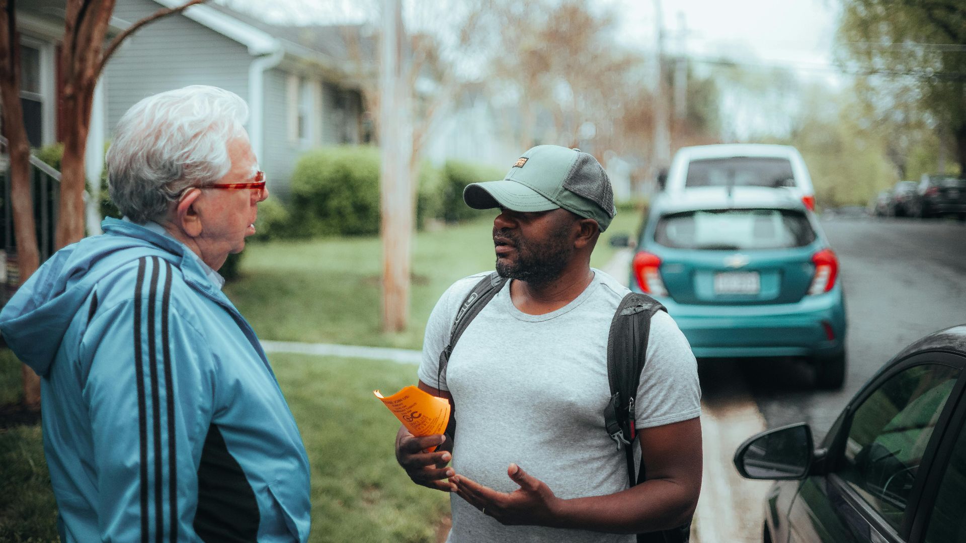 Two men engaged in conversation on a suburban sidewalk with parked cars.