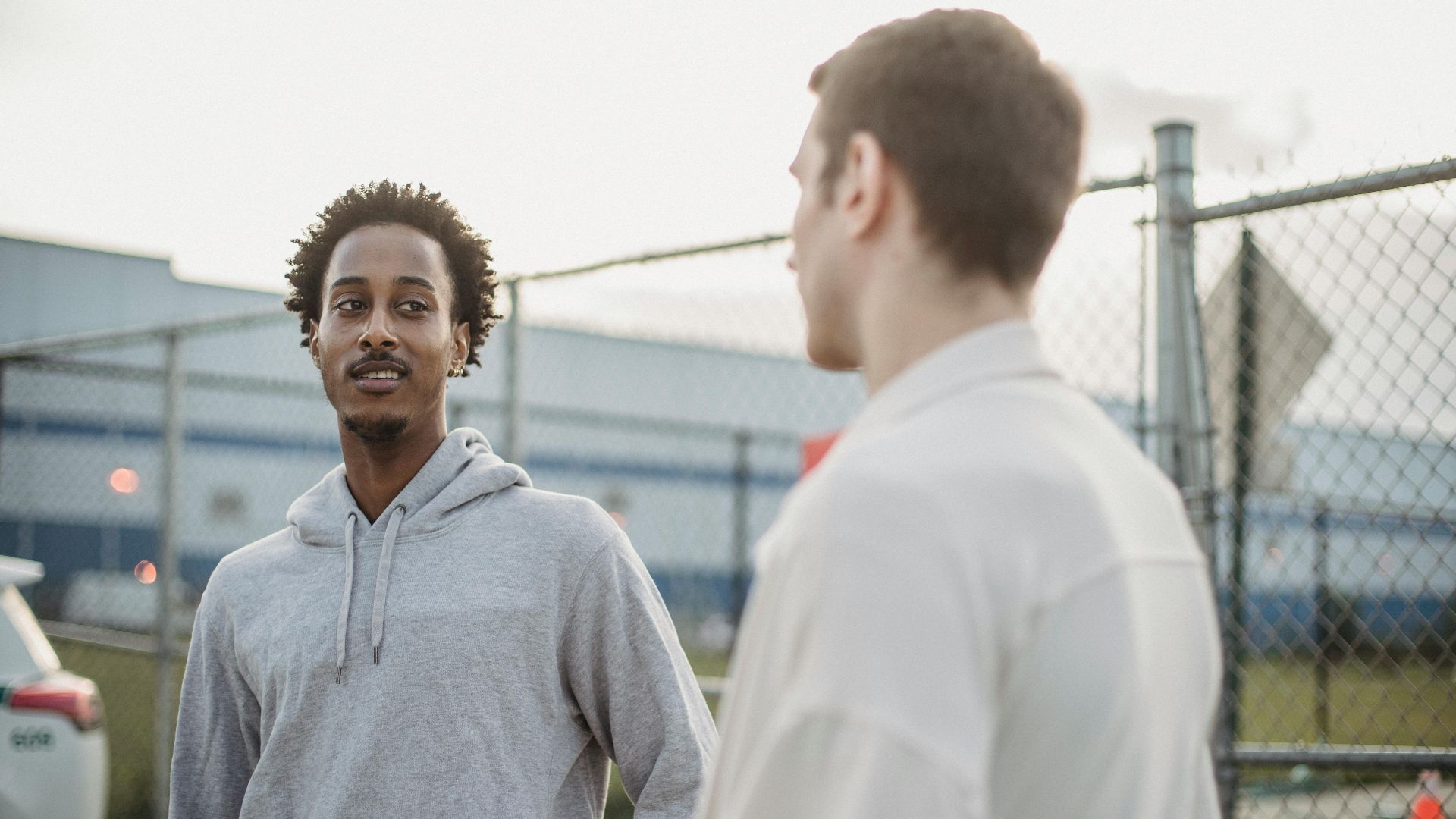 African American male standing with friend against metal grid near car on parking lot