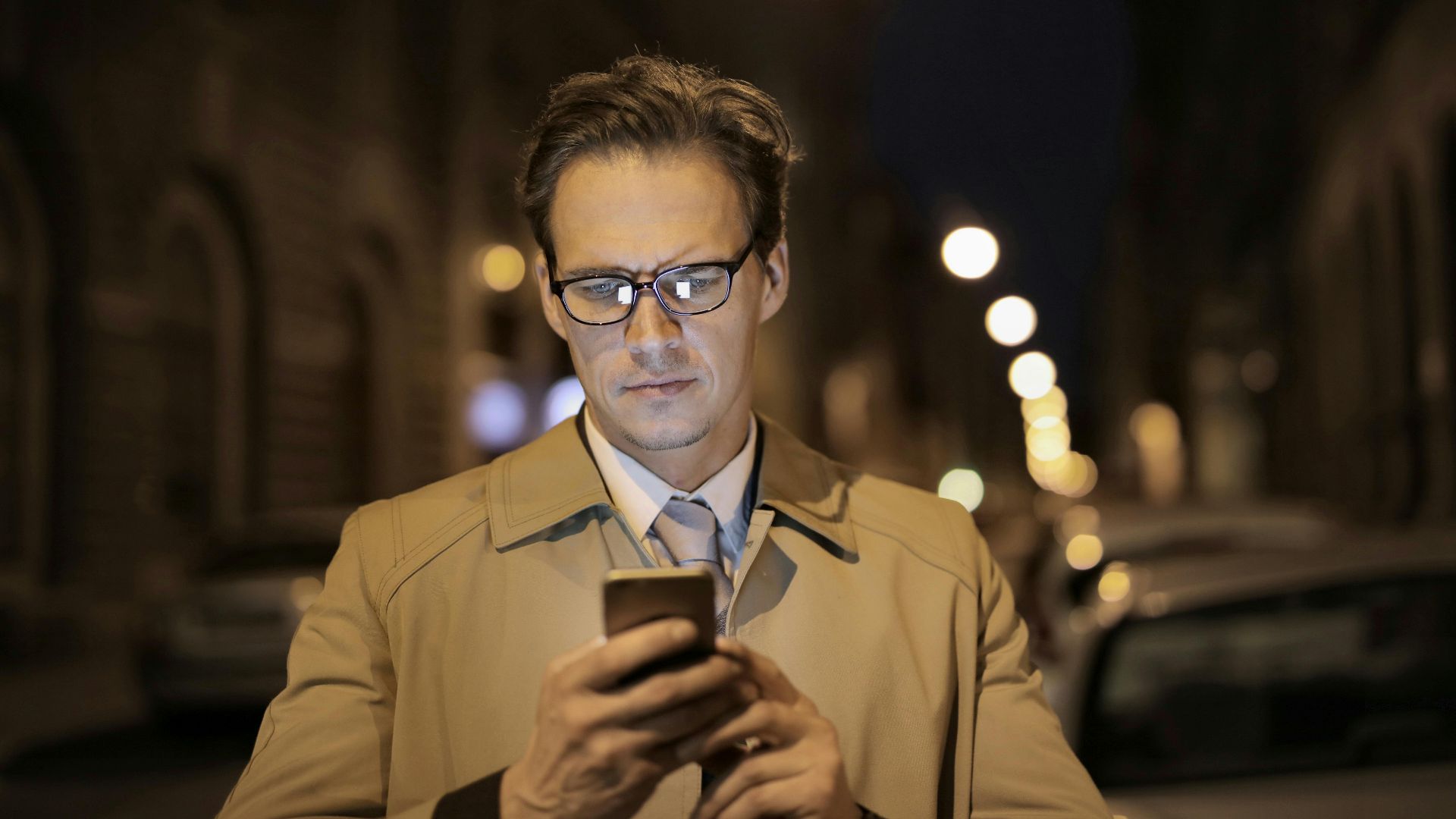 Serious formally dressed man in glasses messaging on phone while standing on city street at night