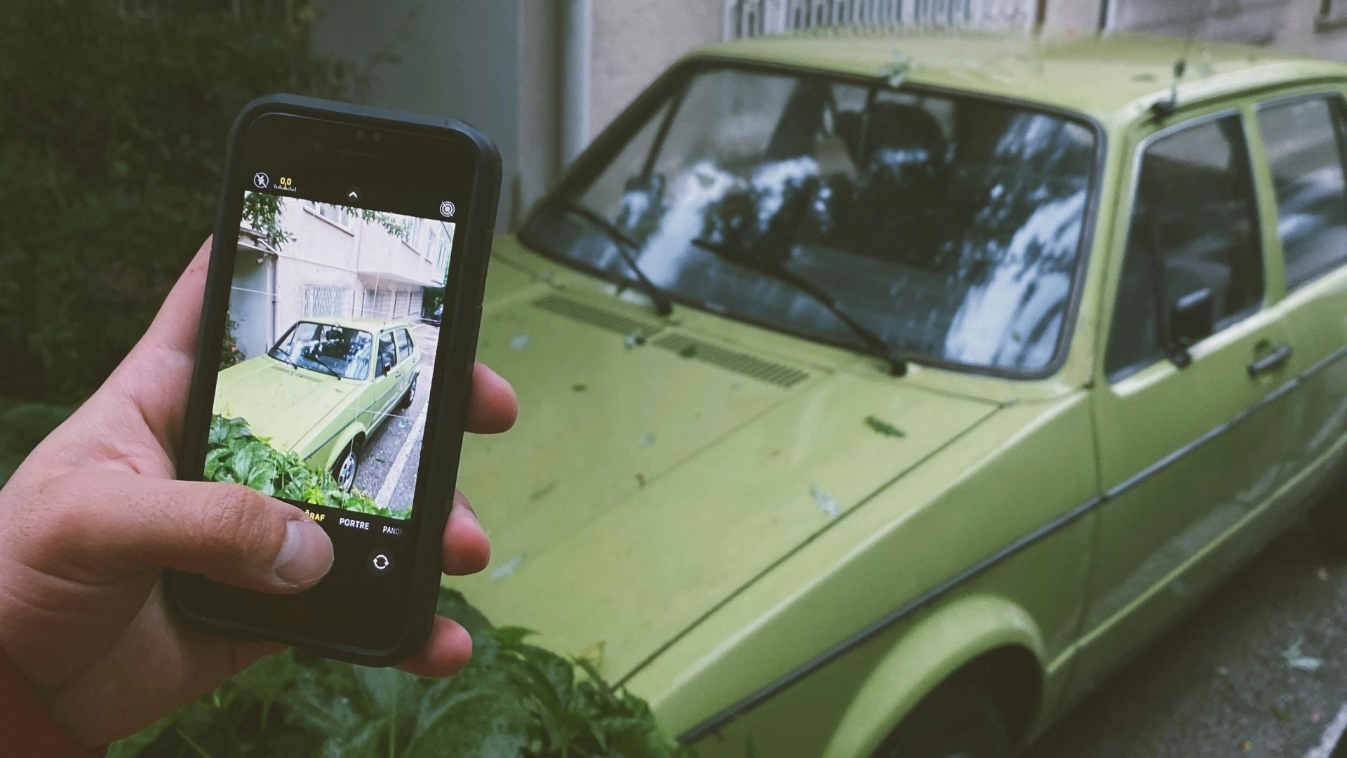 A hand holding a smartphone captures an image of a vintage green car parked by a residential building.
