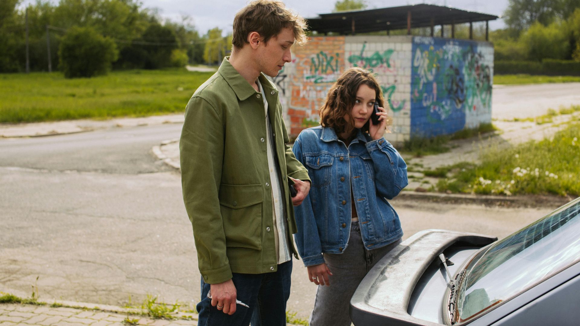 A young couple stands by their car, possibly facing a breakdown.