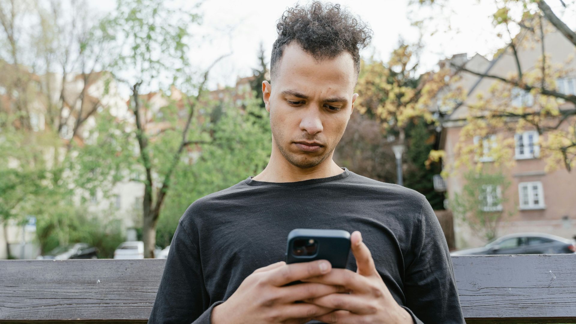 Casual young man sitting on a bench, texting on his smartphone in a park setting.