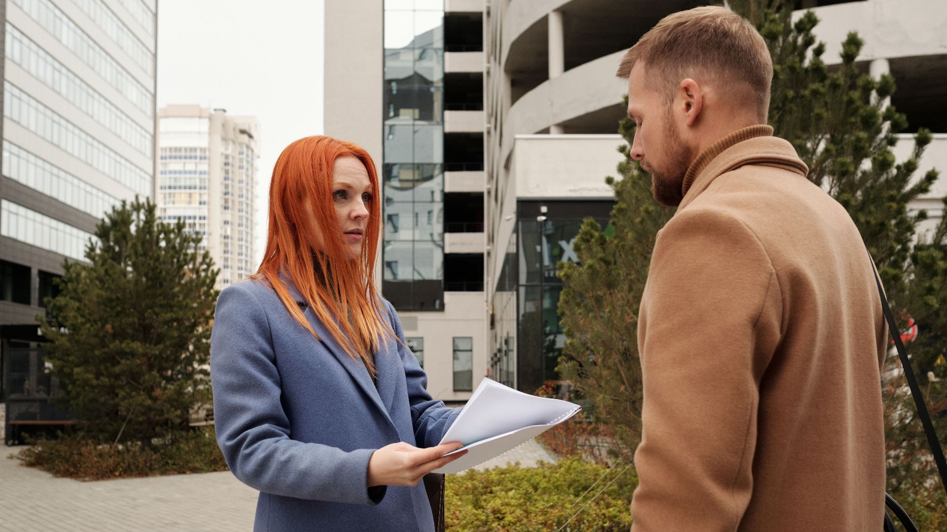 Man and woman in corporate attire engaging in conversation outside urban office complex.