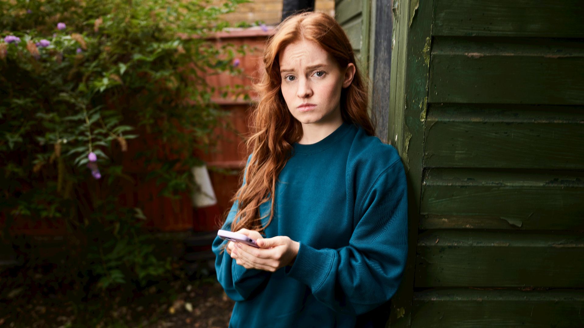 A young woman stands outdoors in a garden holding a smartphone.
