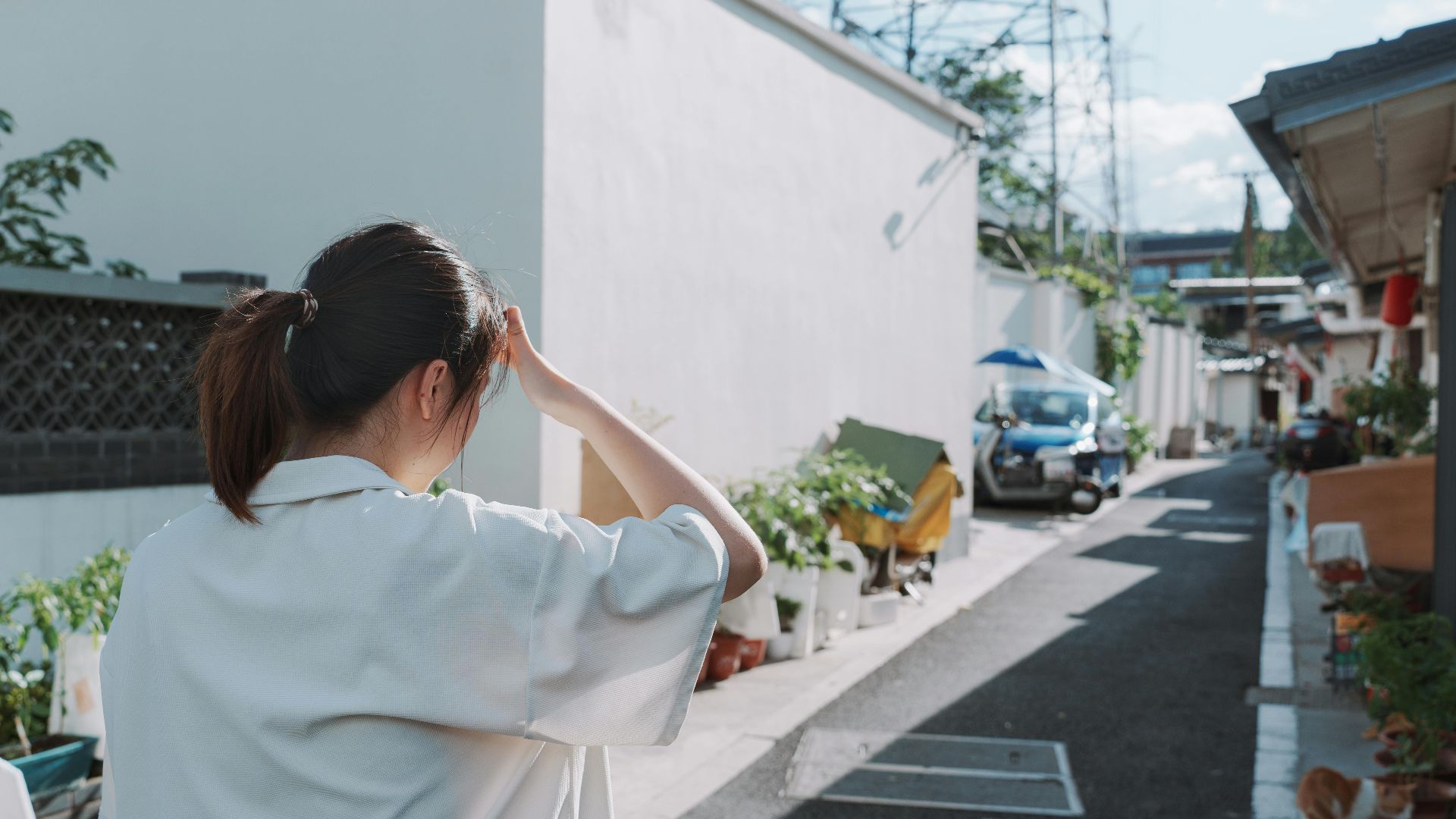 A woman walks through a sunlit alley in an urban area, shielding her eyes.