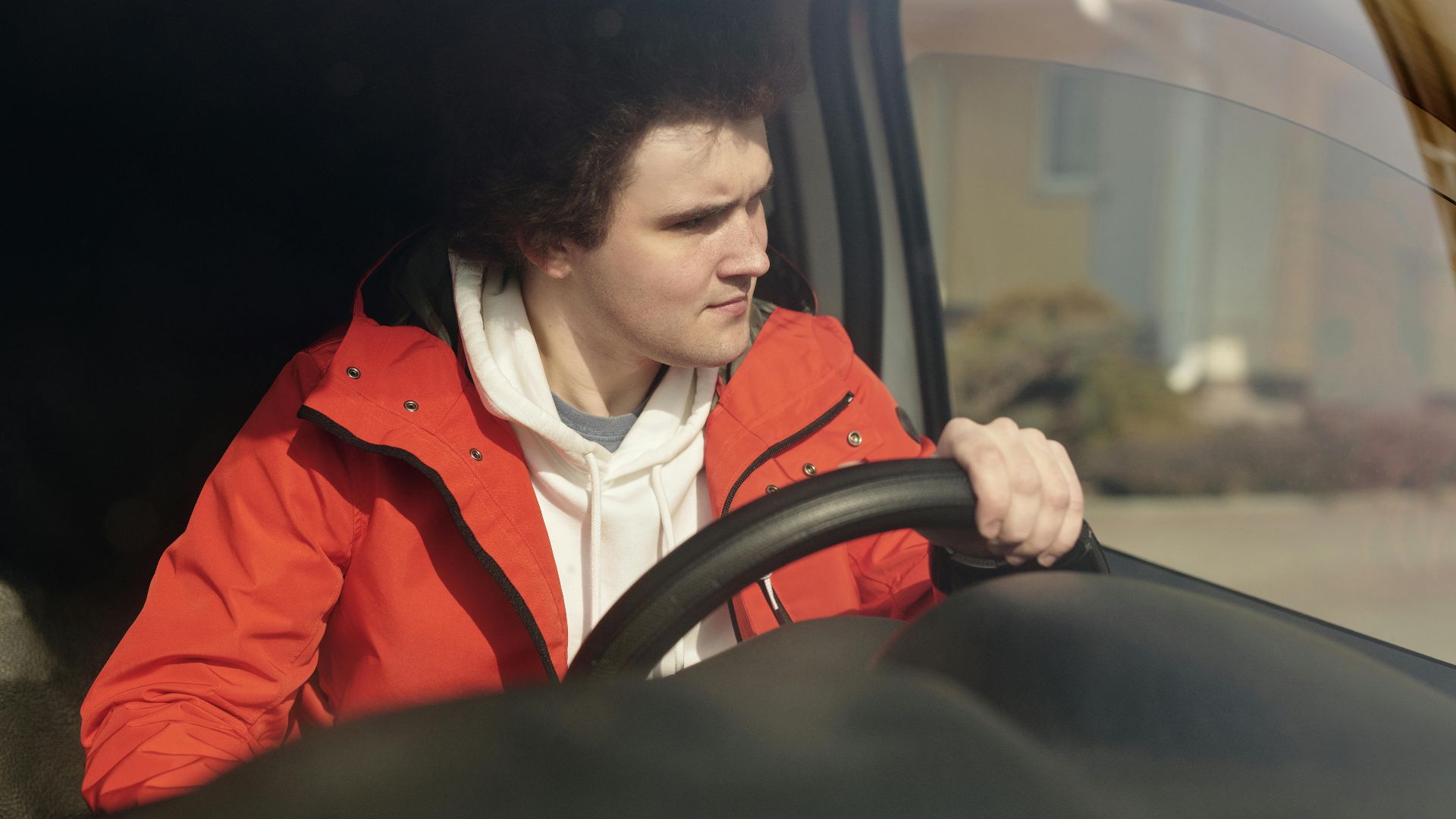 A young man in a red jacket steering a car with focus, enjoying a peaceful drive.