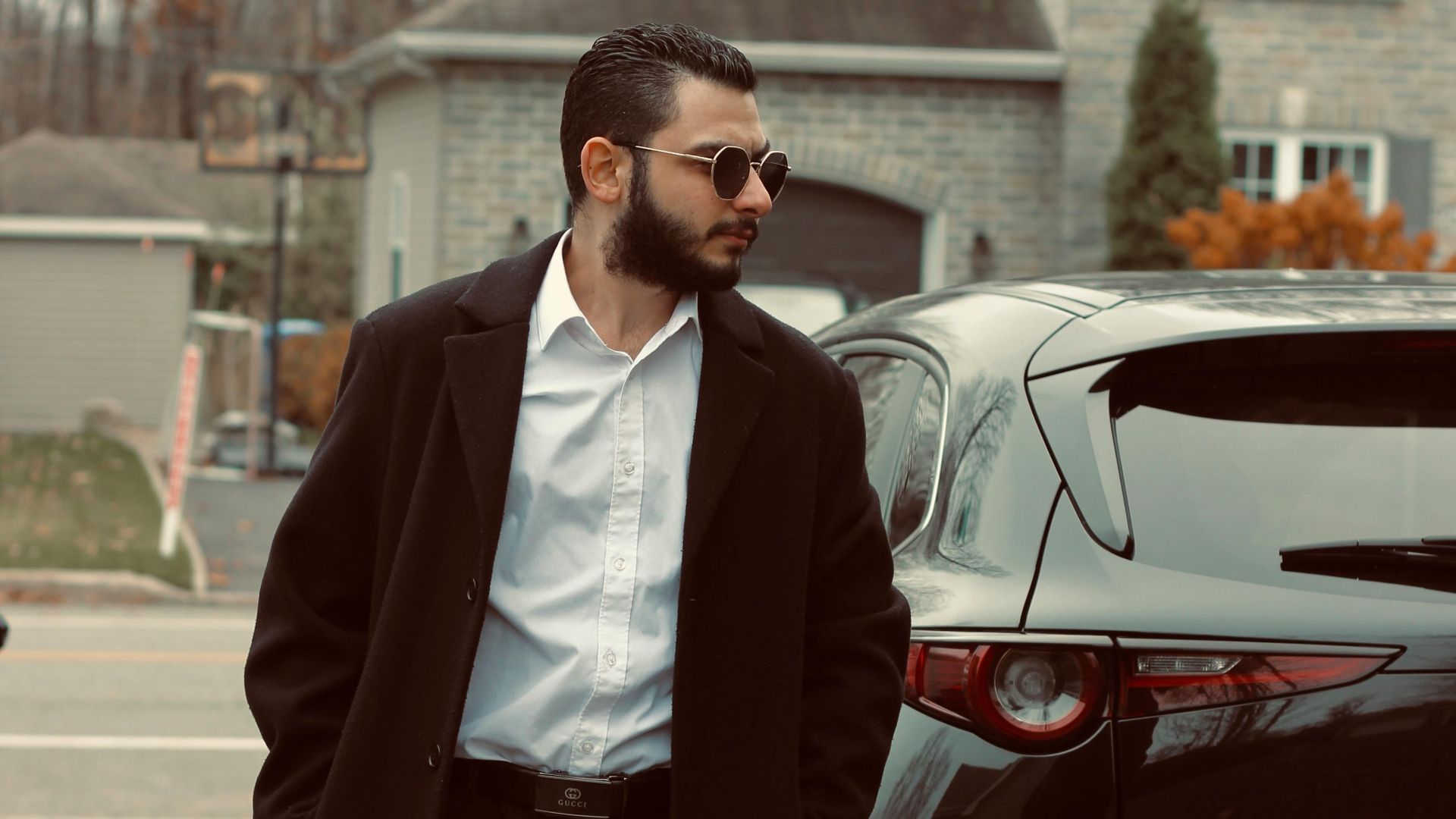 Fashionable man in coat and sunglasses beside car in Canadian neighborhood.