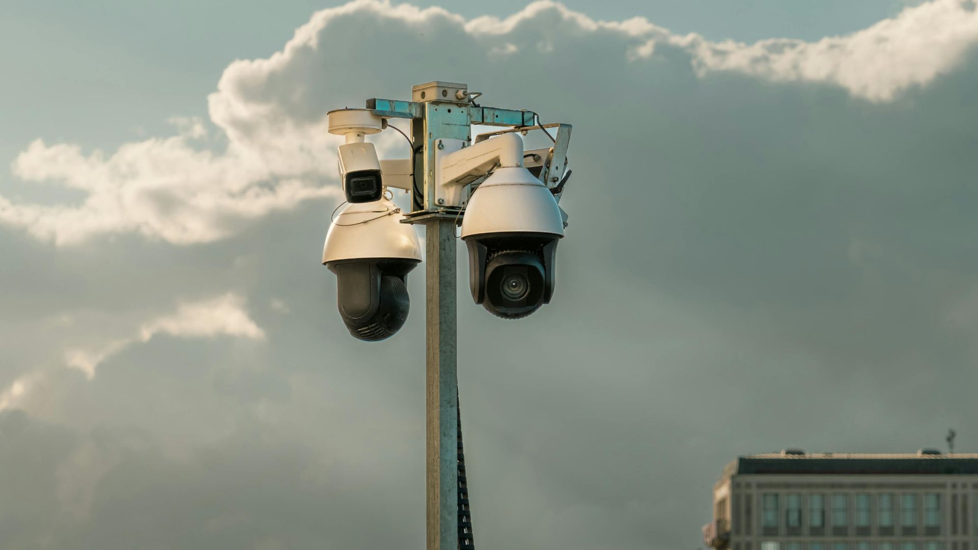Two modern surveillance cameras mounted on a pole in an urban setting under a cloudy sky.