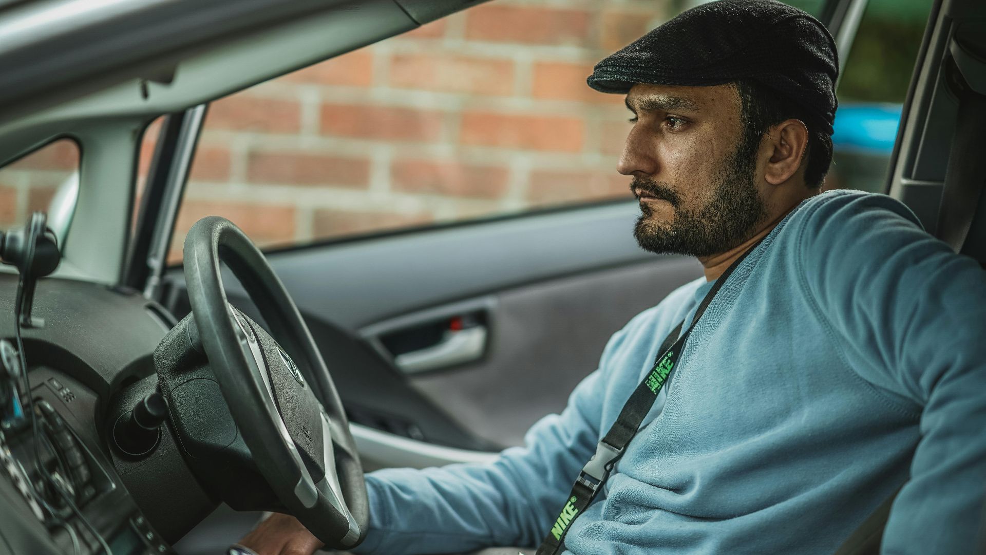 A bearded man wearing a hat and blue shirt sits thoughtfully behind the wheel of a car.