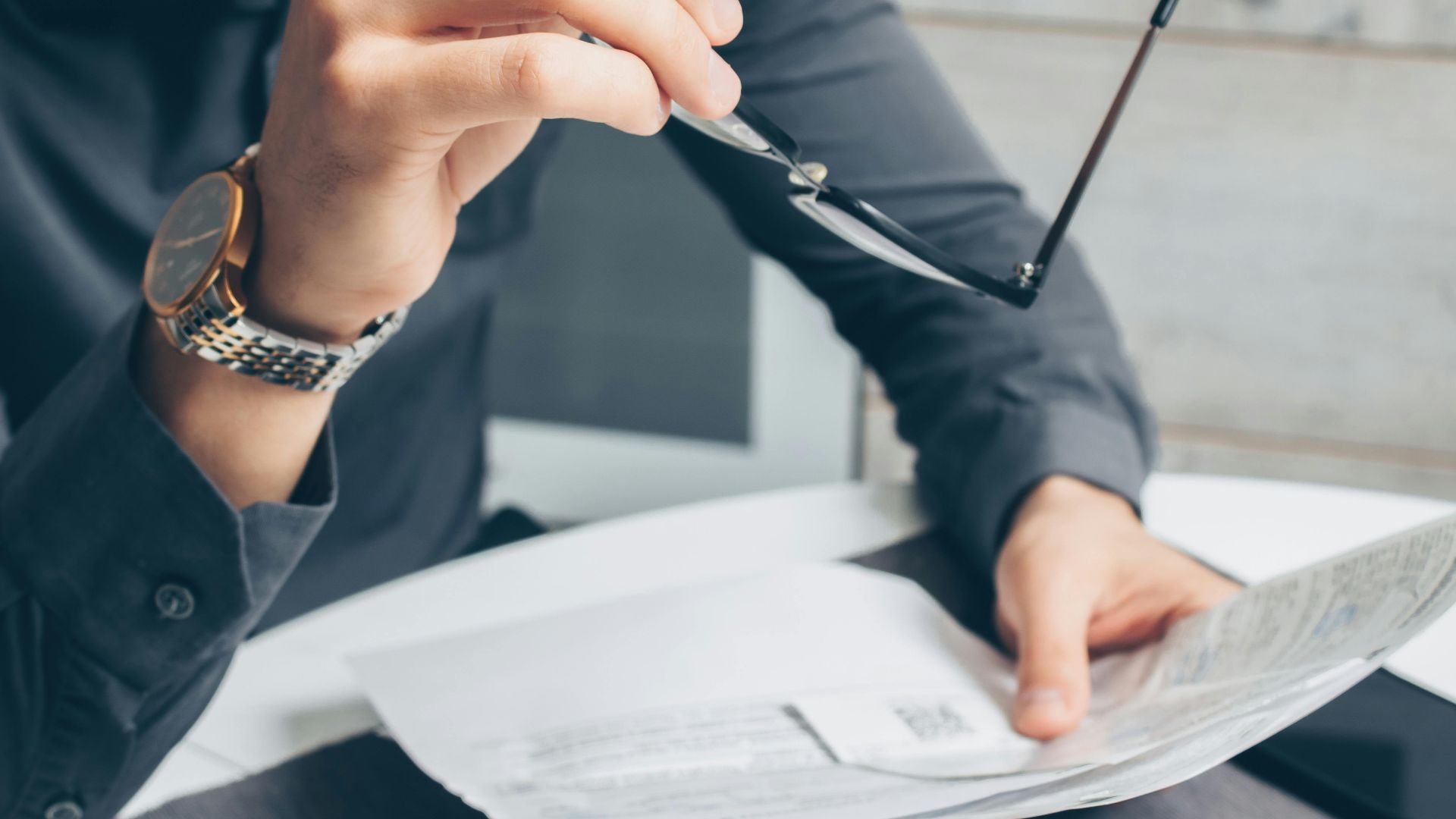 Man in formal attire reviewing paperwork, holding glasses. Business setting.