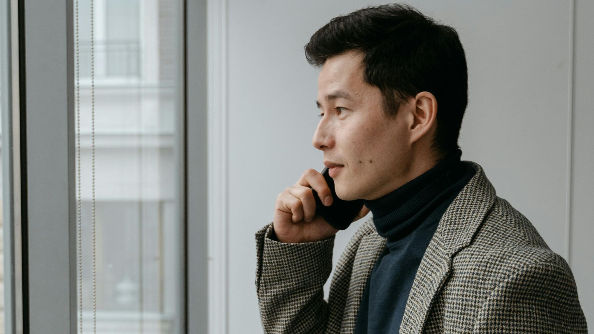 Confident businessman in a gray blazer talking on phone by office window.