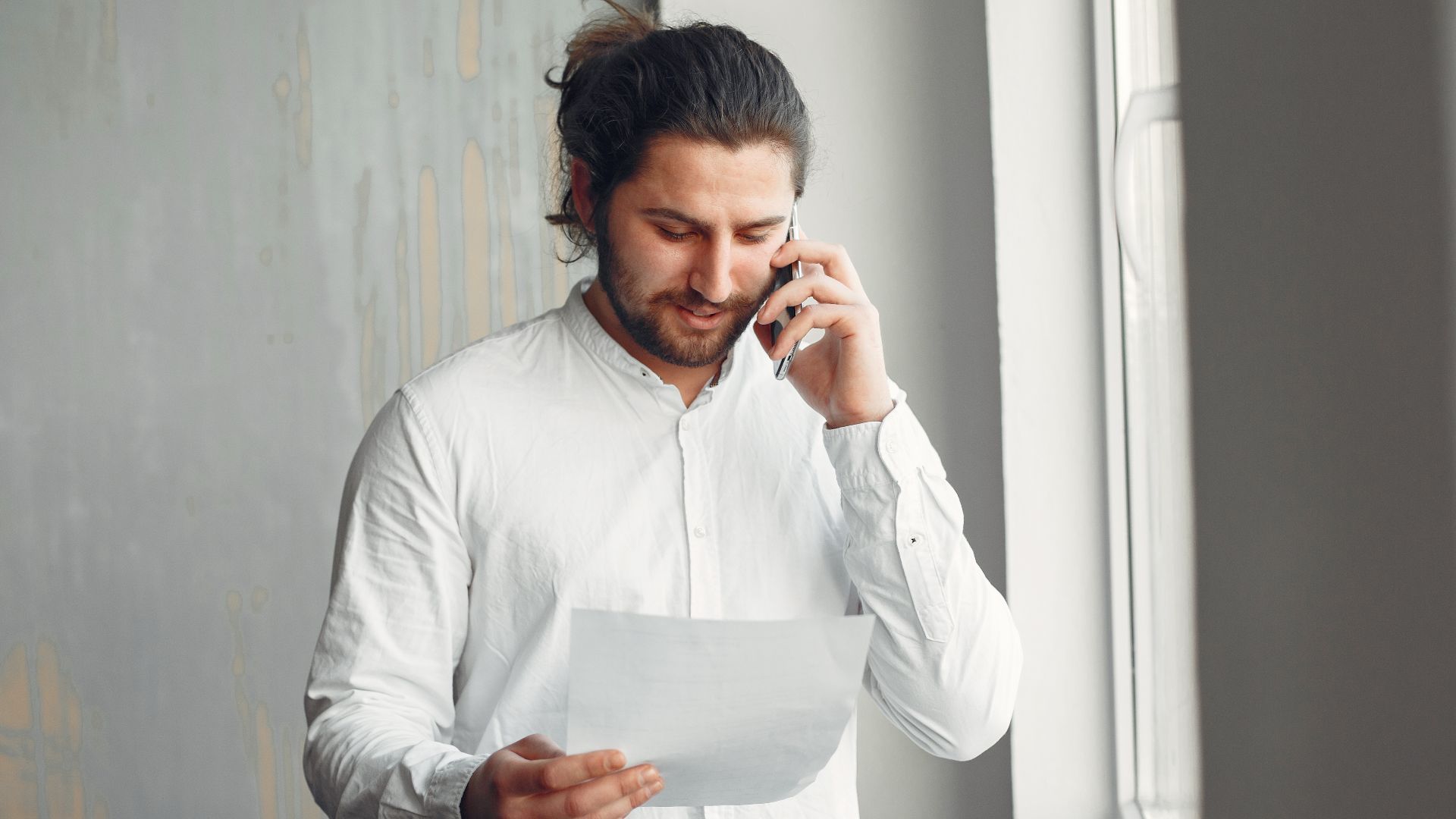 A young man in casual attire talks on the phone while holding a document indoors.