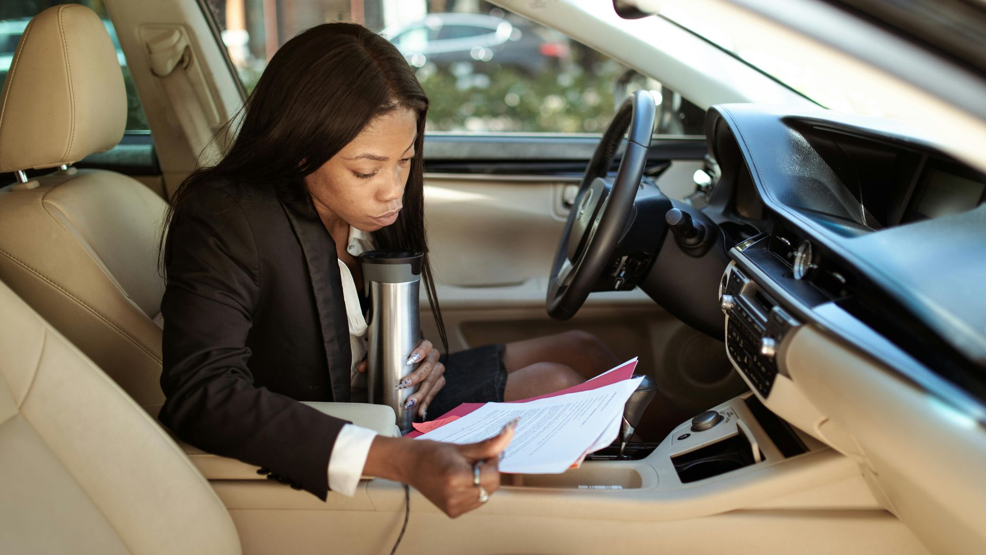 A focused businesswoman reads documents in her car, multitasking with a tumbler.