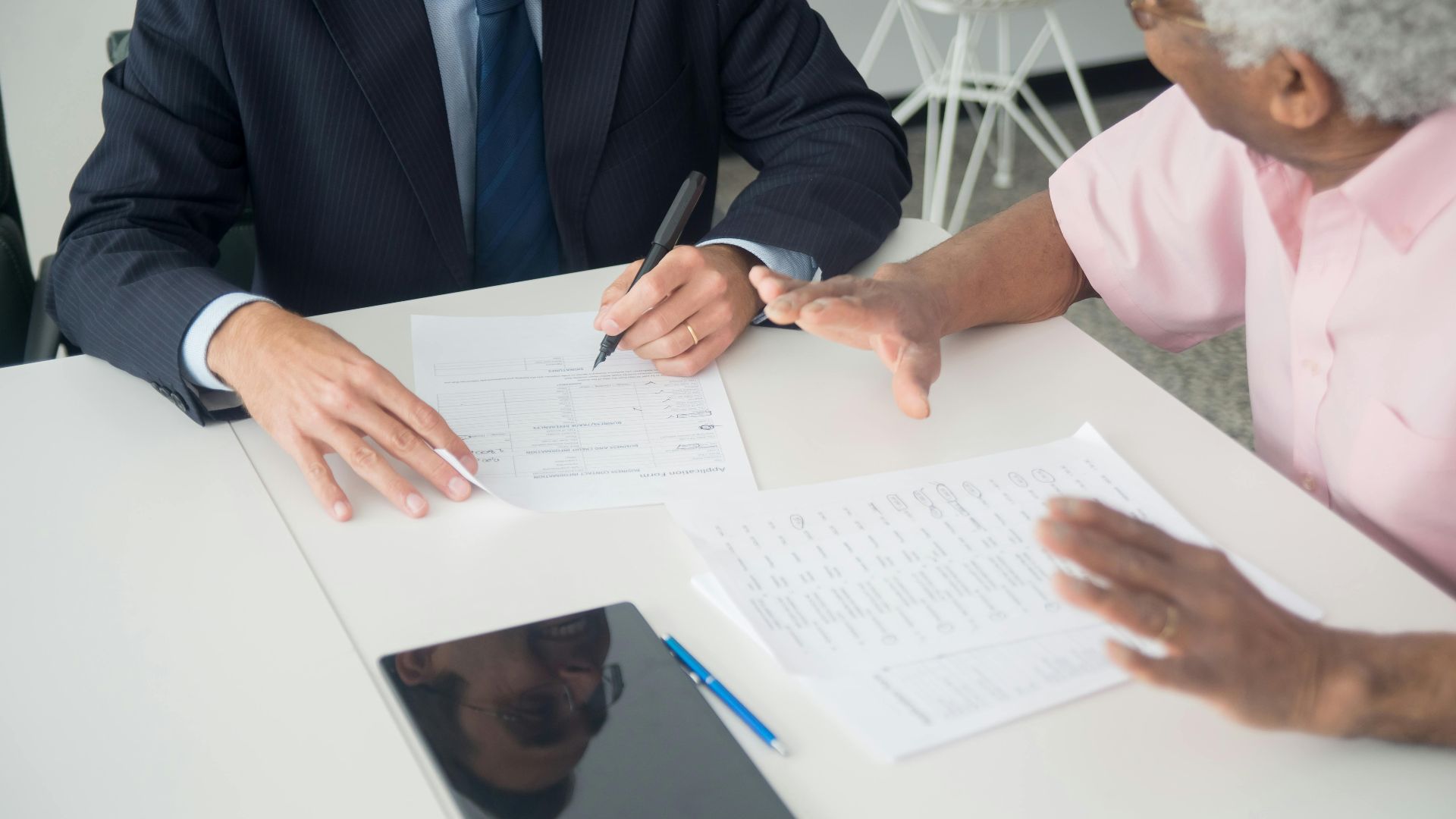 A business meeting between an elderly client and a consultant discussing documents at an office table.
