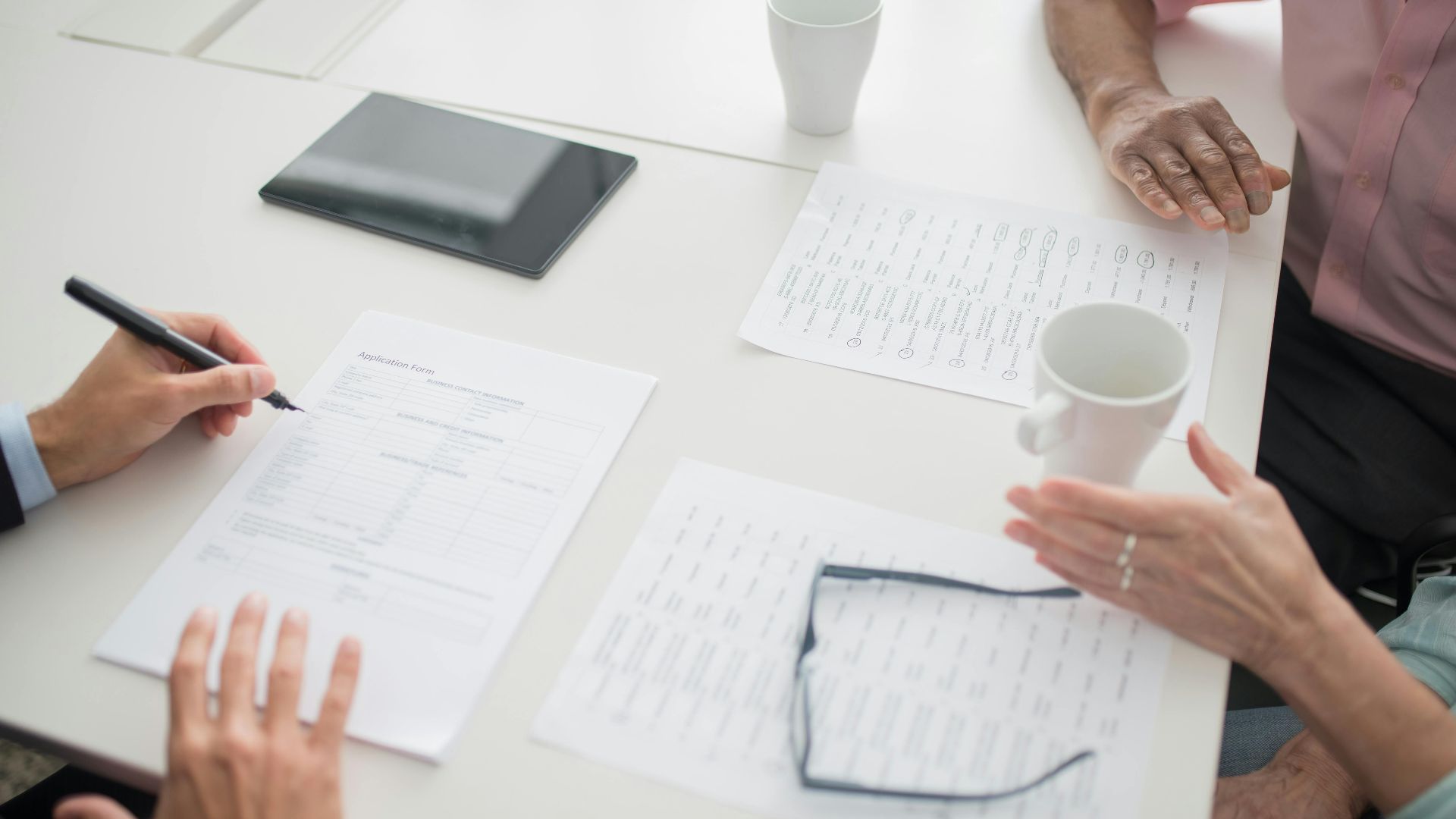 Hands writing on documents in a business meeting setup with coffee cups and tablet.