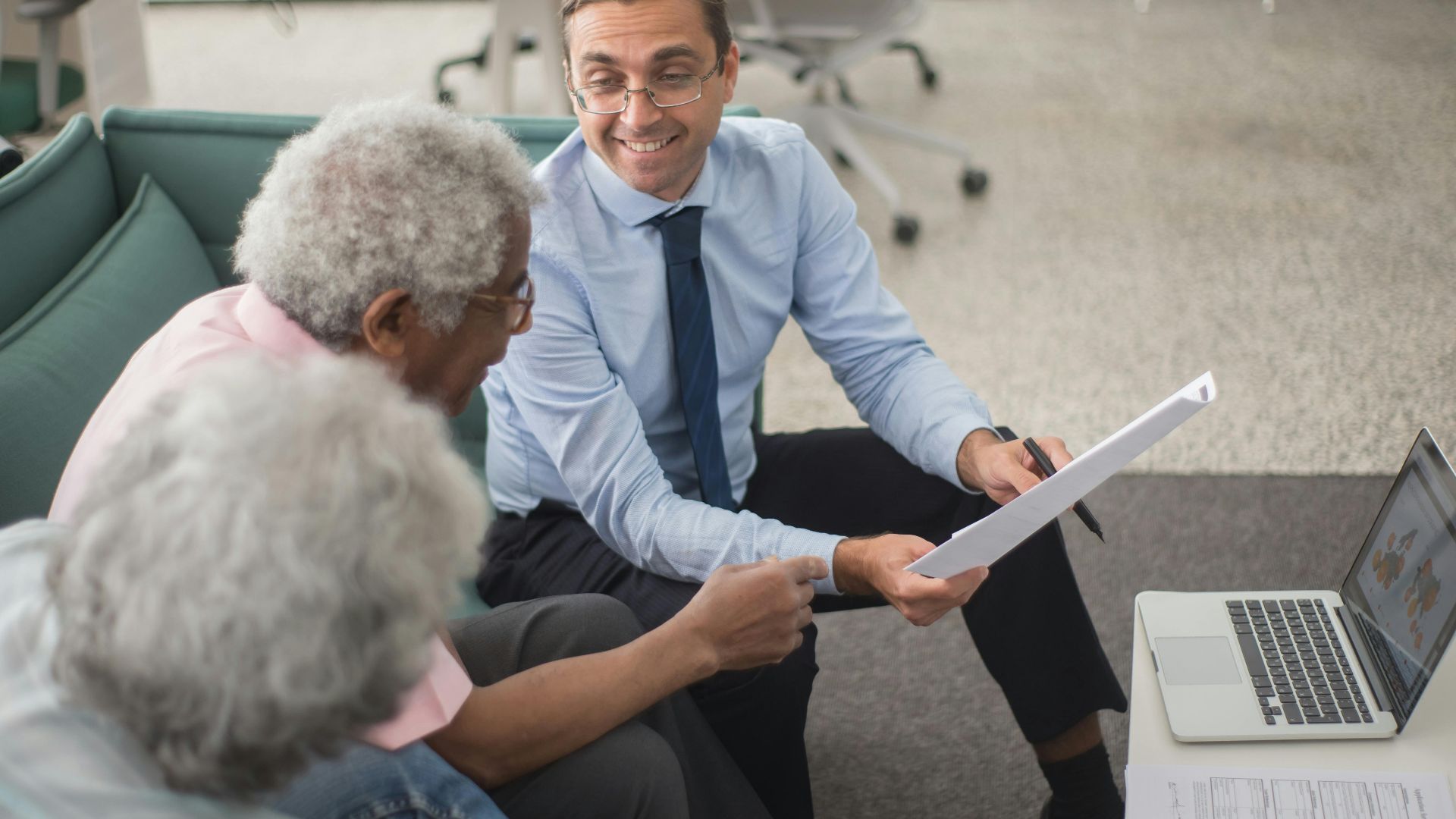 Consultant discussing financial plans with senior clients in a modern office setting, using documents and a laptop.