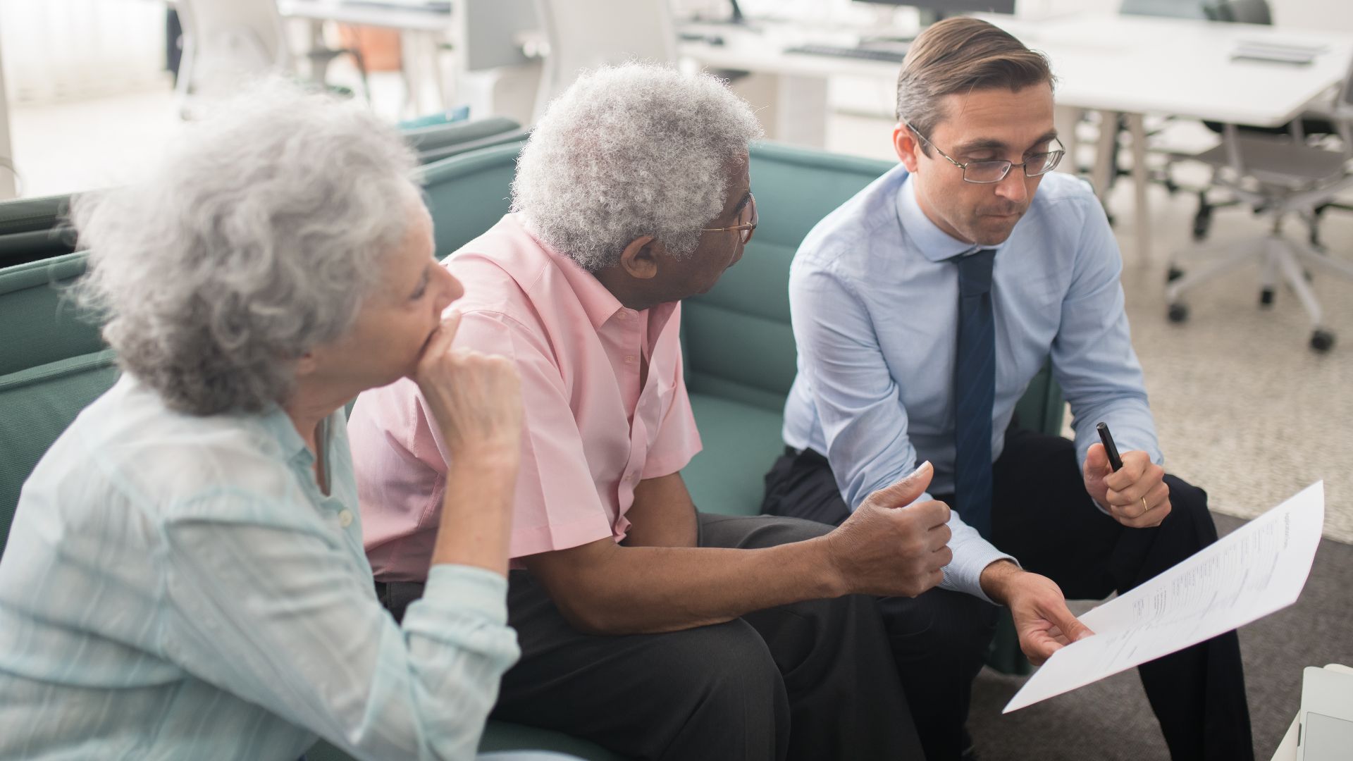 Business professional consults elderly clients in an office setting. Collaborative discussion, paperwork visible.