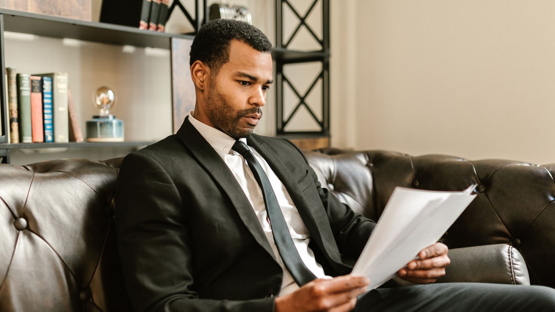 Businessman in suit reading papers on a leather sofa in a modern office setting.
