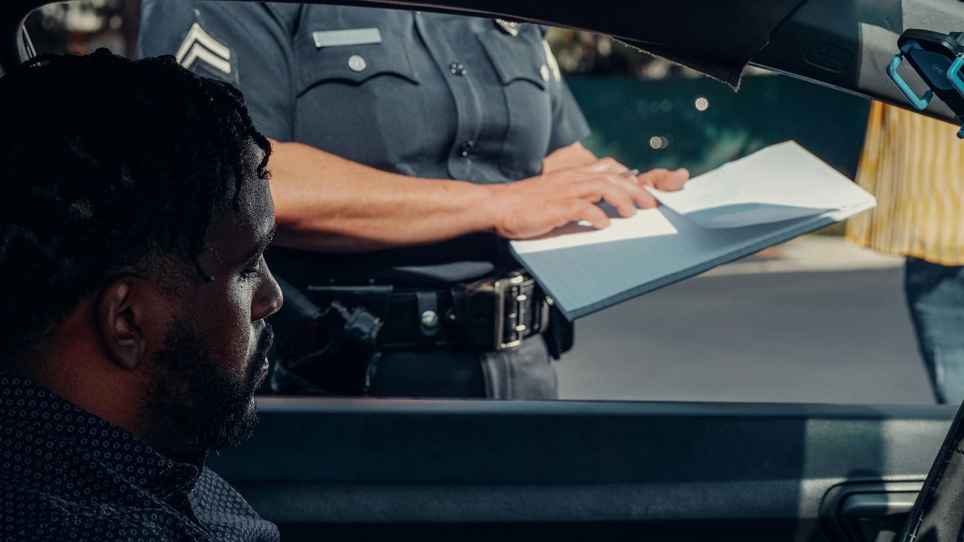 A police officer issues a traffic ticket to a man in a car. Close-up view of the interaction through the window.