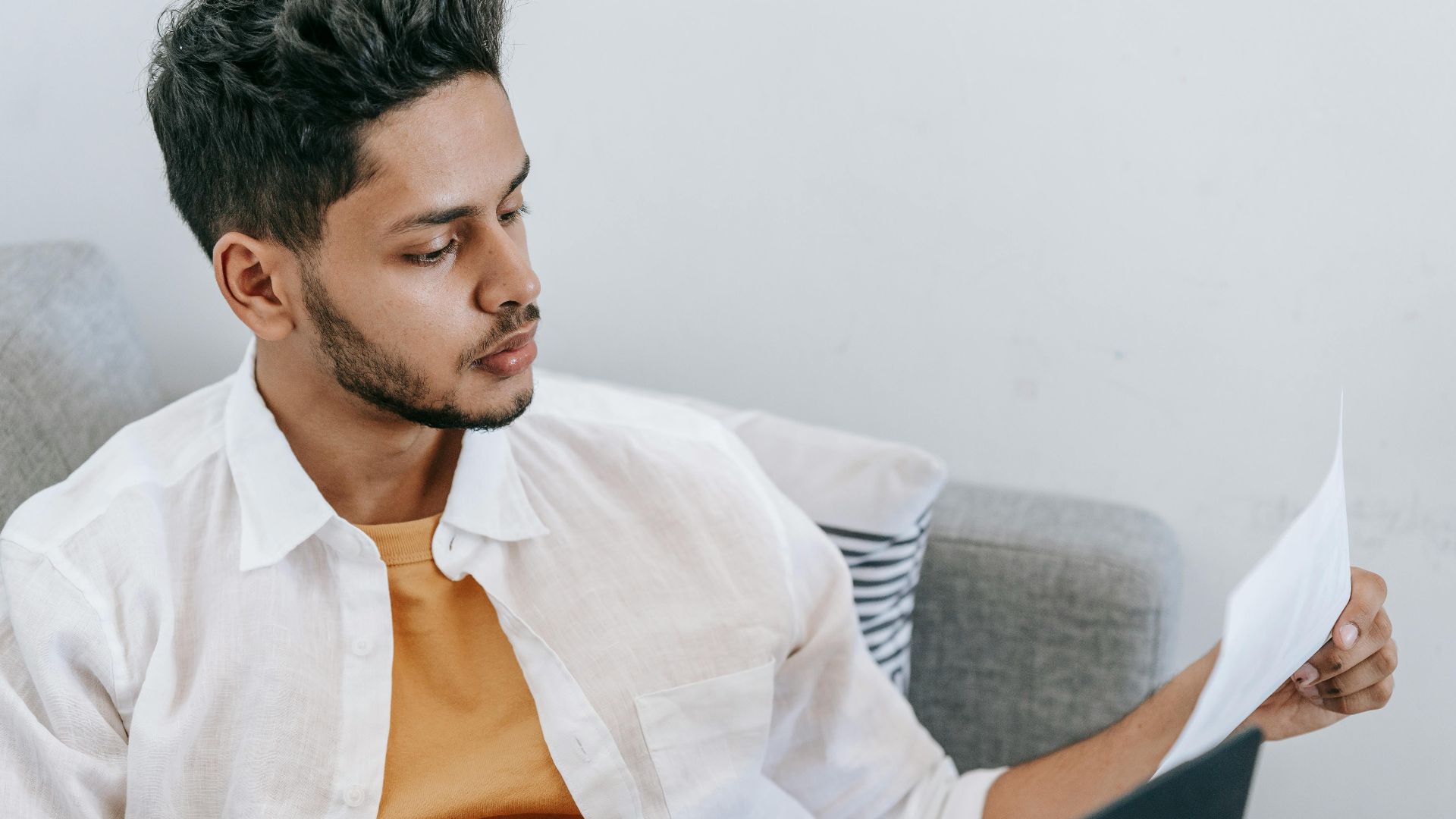 Concentrated young ethnic male wearing casual outfit reading document and browsing netbook while resting on cozy couch
