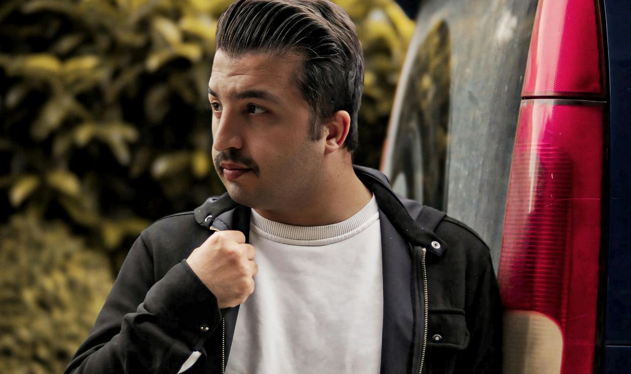 A man in casual attire poses next to a car, set against a backdrop of green foliage outdoors.