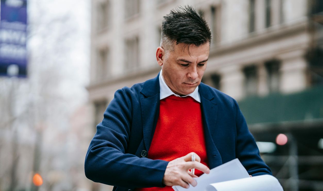 Man in a stylish outfit examines paperwork outside a city building, focused and pensive