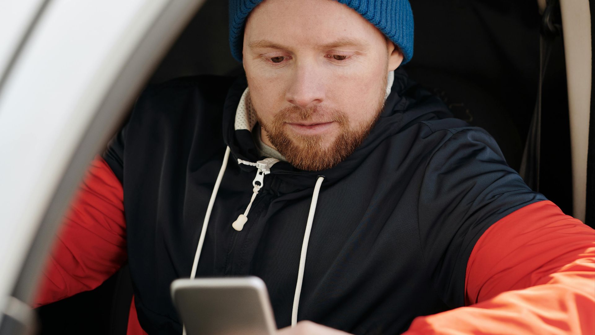 A man with a beanie uses a smartphone in a parked car.