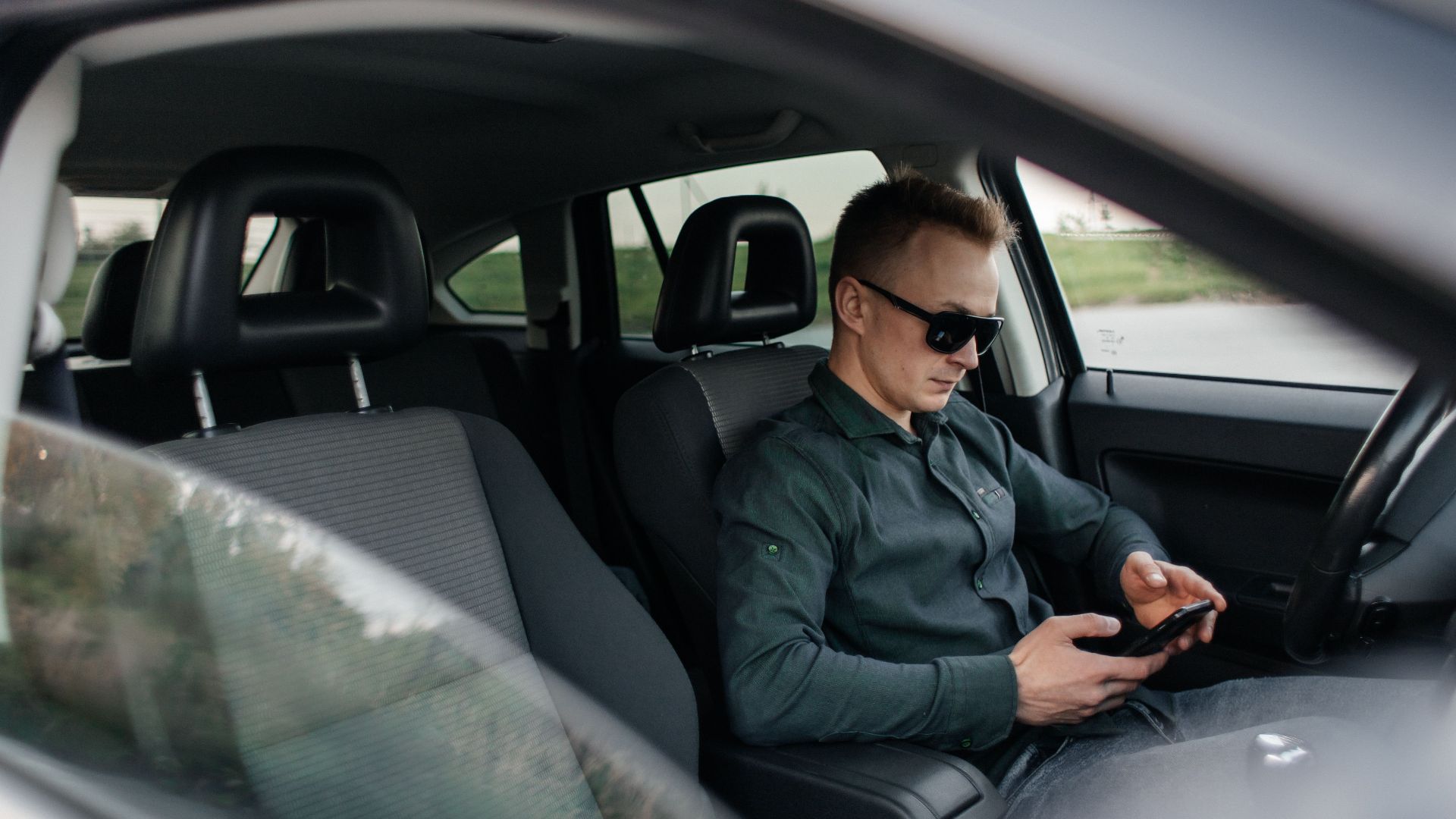Young man using smartphone inside a parked car, wearing sunglasses.