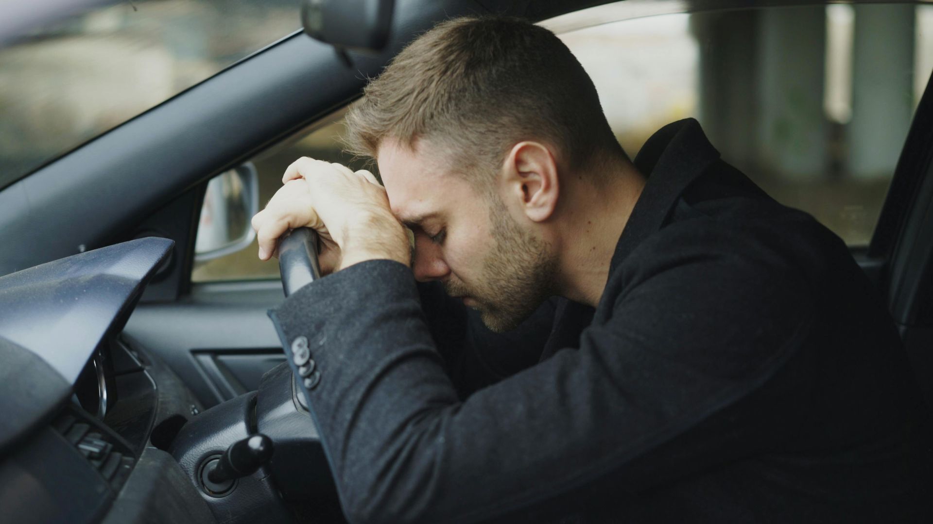 A man showing stress while sitting in a car, head on steering wheel.