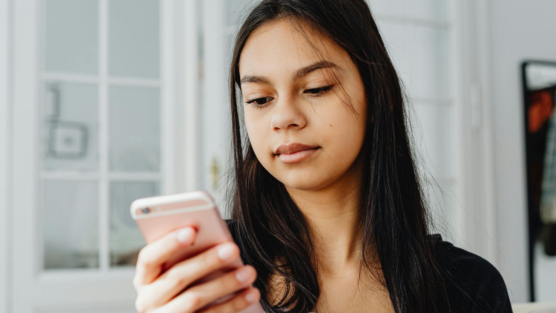 Teenage girl using smartphone indoors with a thoughtful expression.