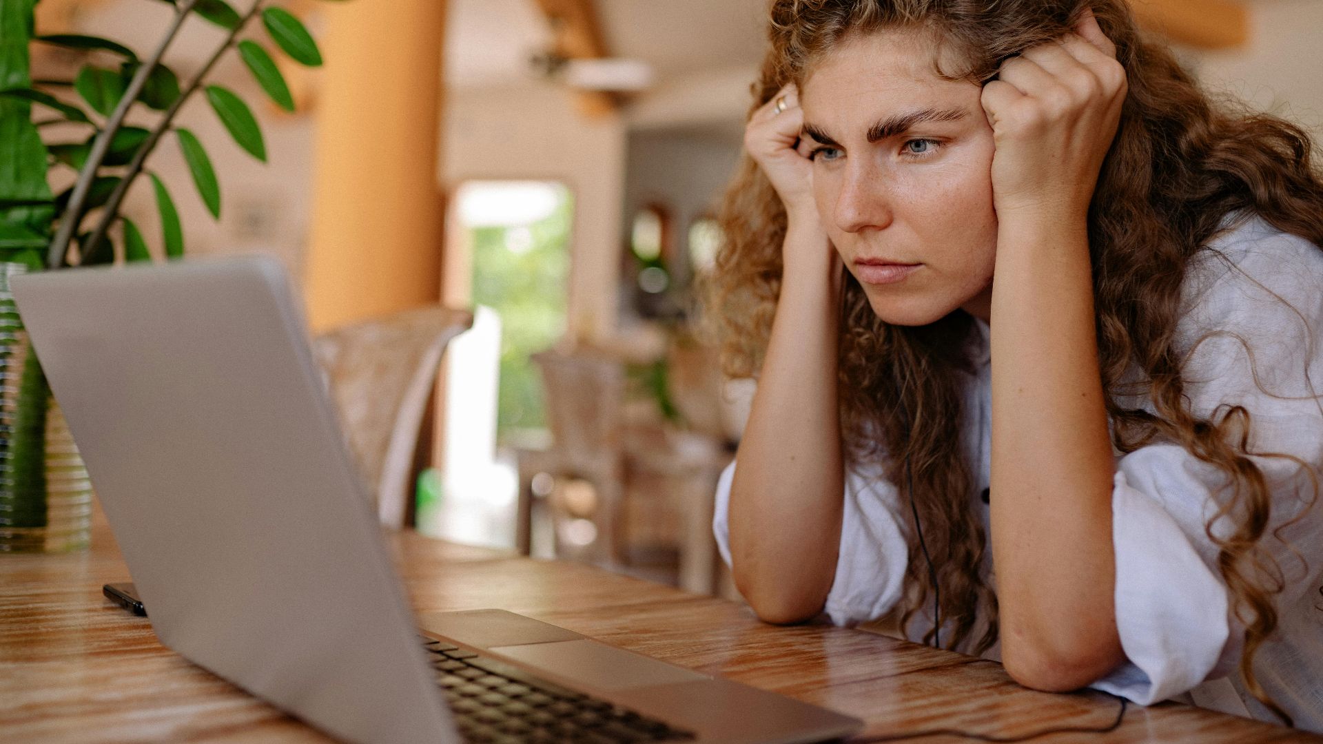 A woman with curly hair intensely focusing on her laptop indoors with plants and natural lighting.