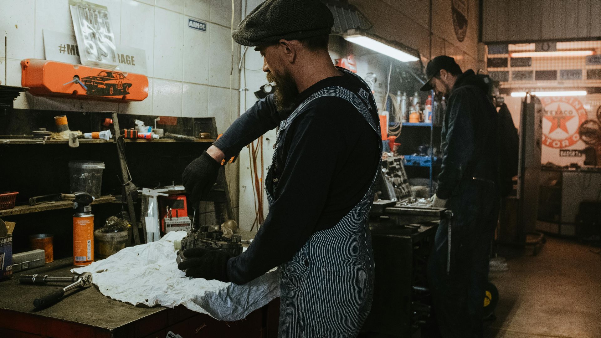 Two auto mechanics engaged in vehicle repair work inside a dimly lit garage.