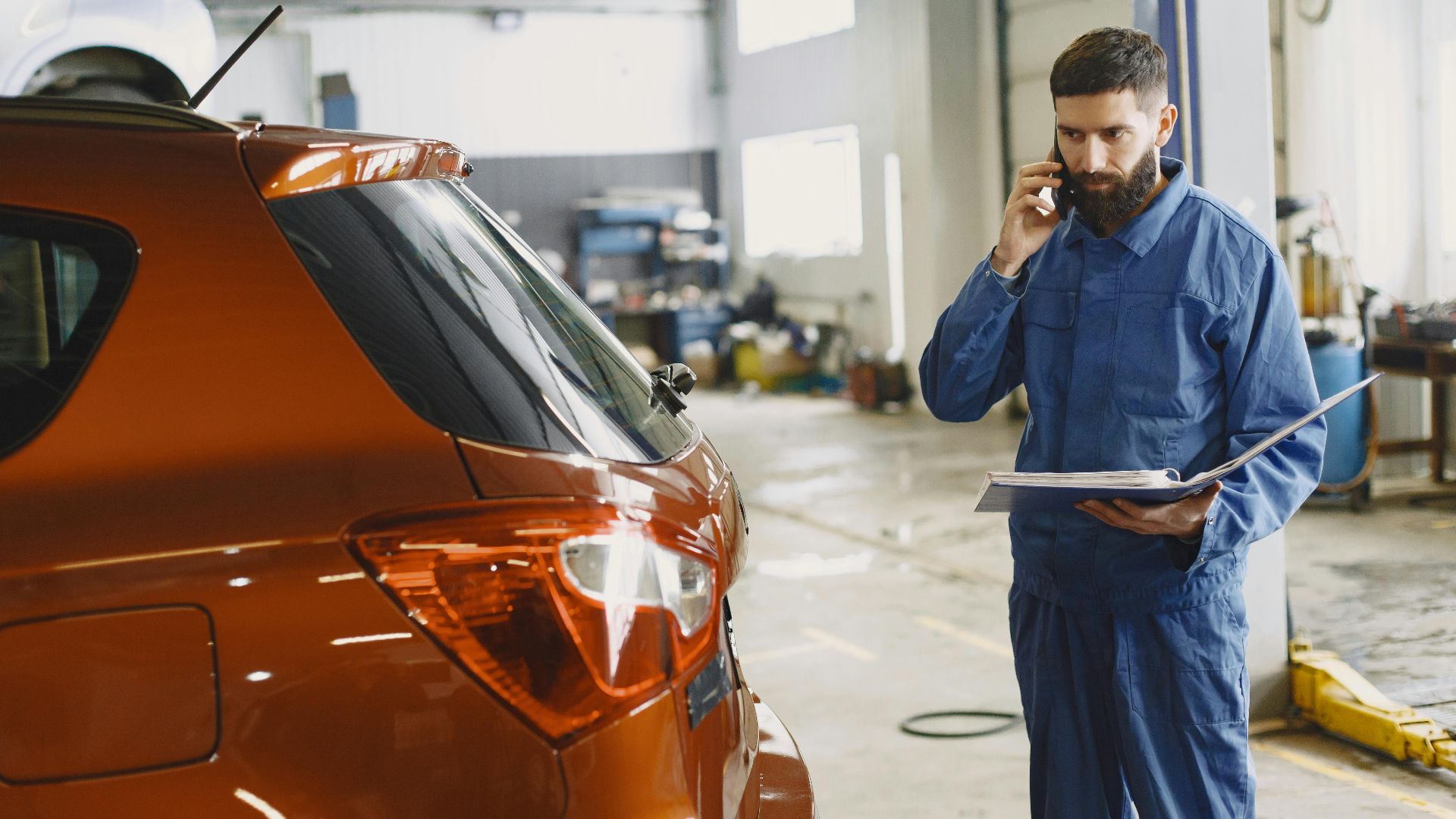 Mechanic in a blue uniform talks on phone beside a red car in an auto repair shop.