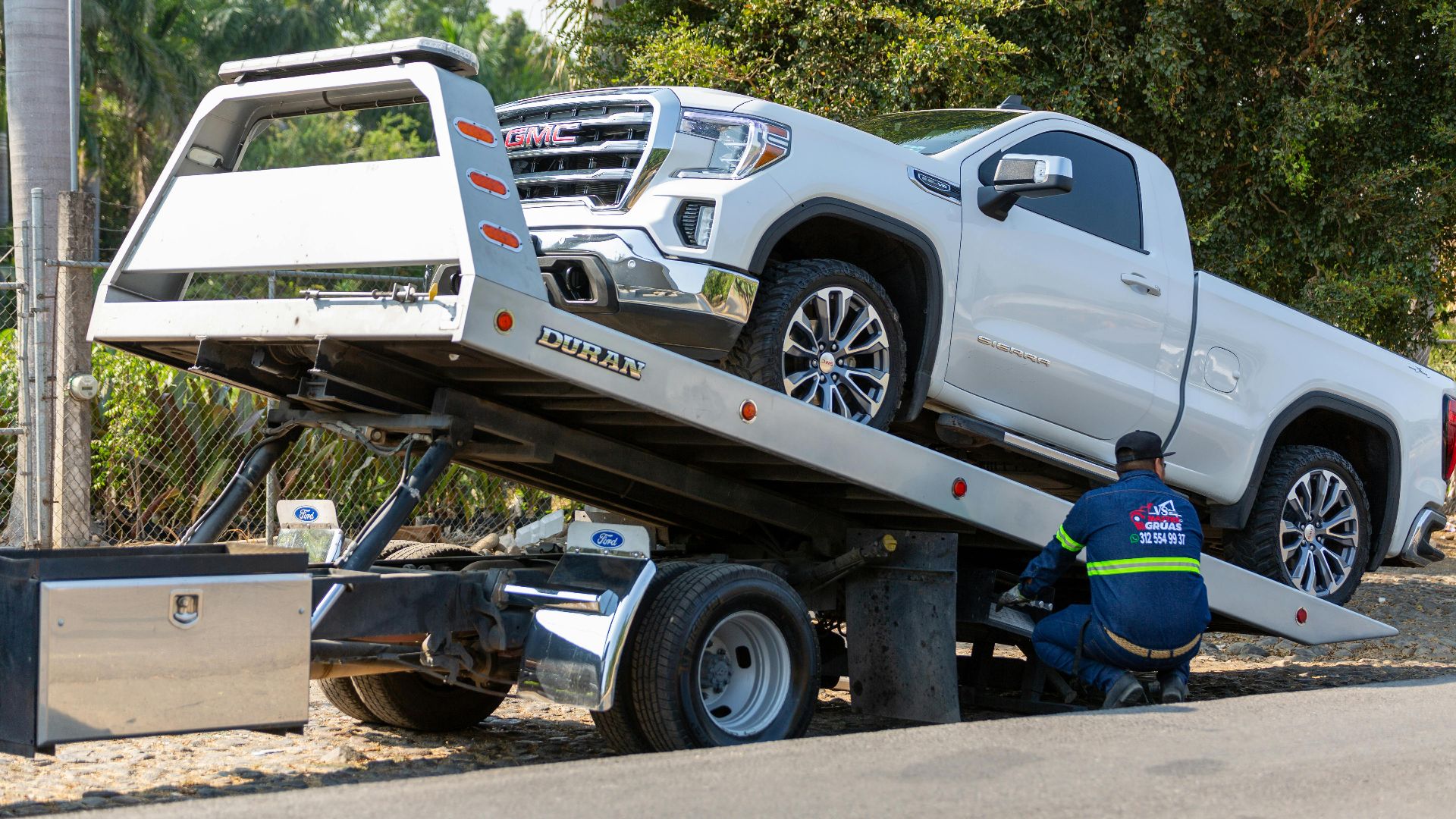 Tow truck operator loading white GMC pickup truck on street in daytime.