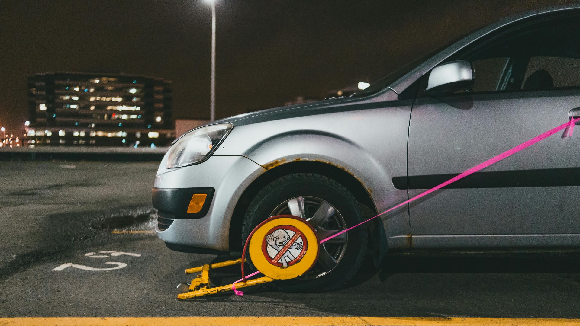 A silver car is immobilized with a wheel clamp in a dimly lit parking lot at night.