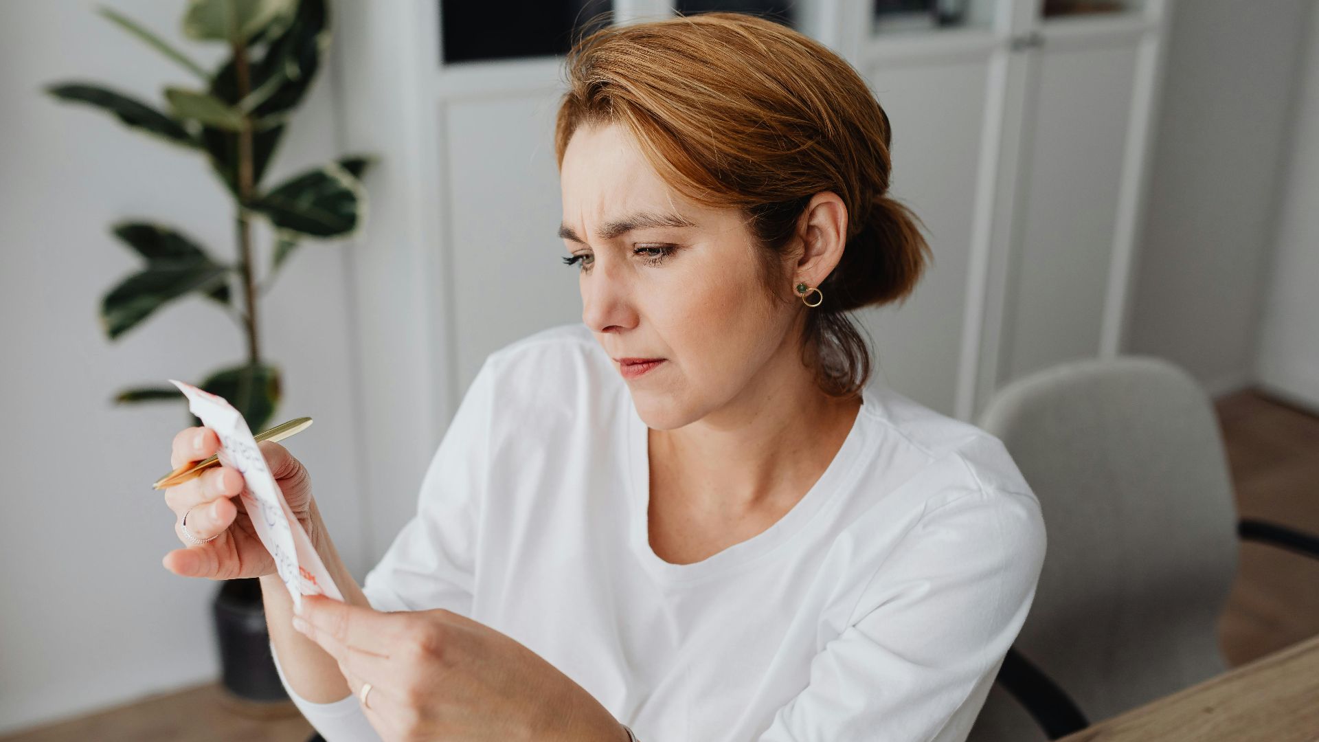 A concerned adult woman examines a receipt while sitting at her office desk, indicating financial scrutiny.